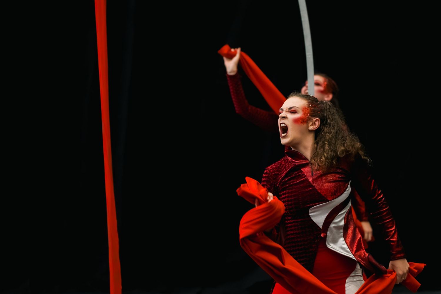 A performer in red costume dances dramatically with flowing red ribbons against a dark stage background.