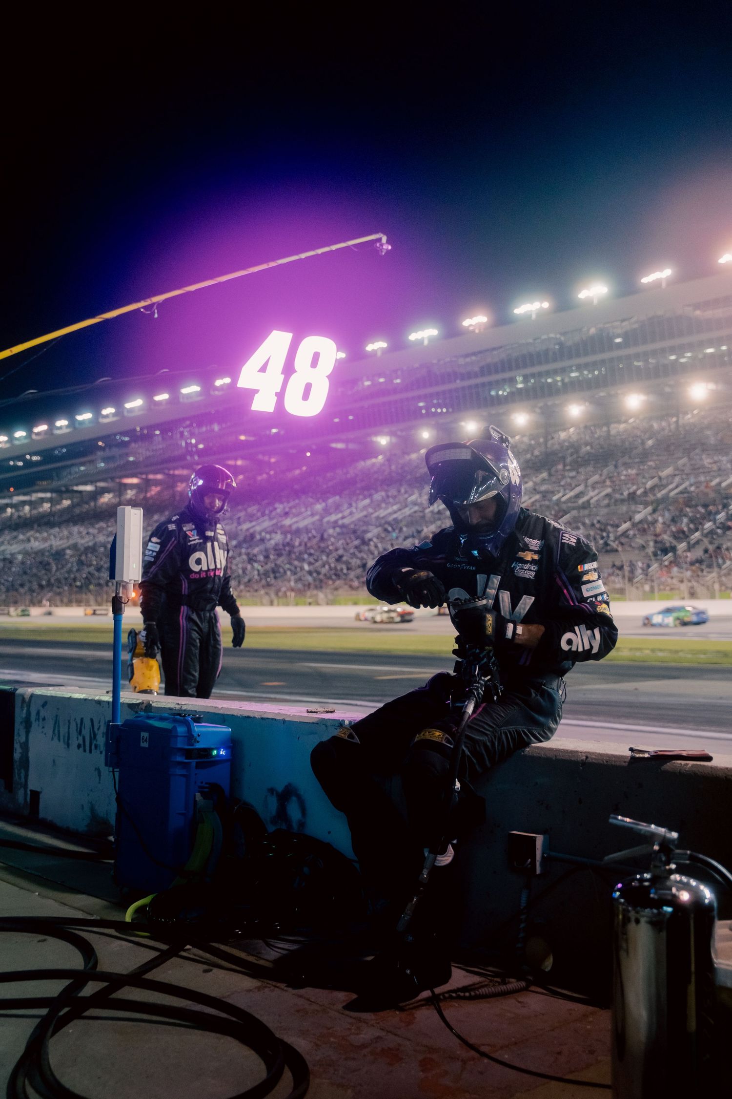 NASCAR pit crew members in Ally-sponsored gear prepare trackside at night, with the iconic #48 car number illuminated above.