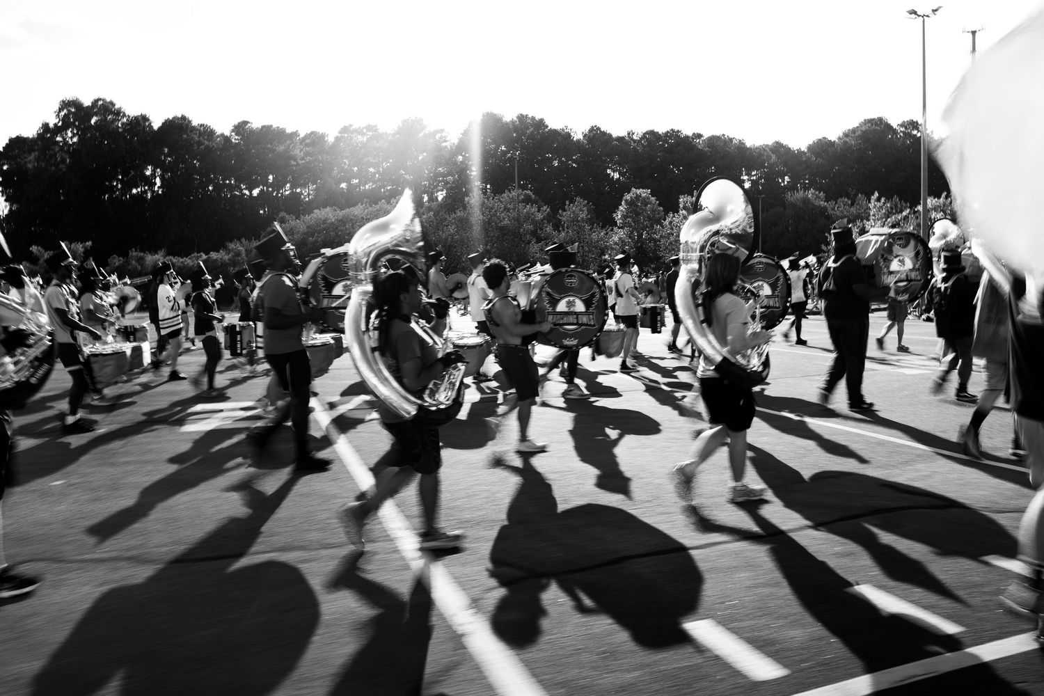 Black and white photo of a marching band performing outdoors in sunlight, casting long shadows on the pavement.