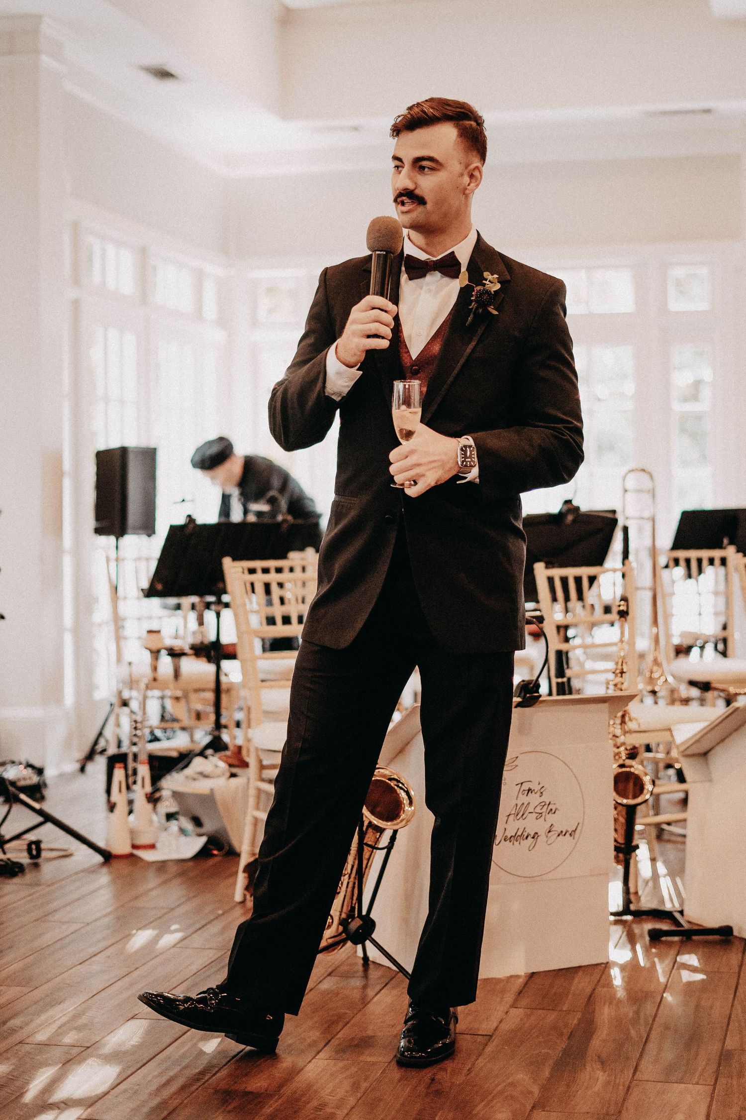 A speaker in a tuxedo holds a microphone while performing at an elegant indoor venue with musical instruments.