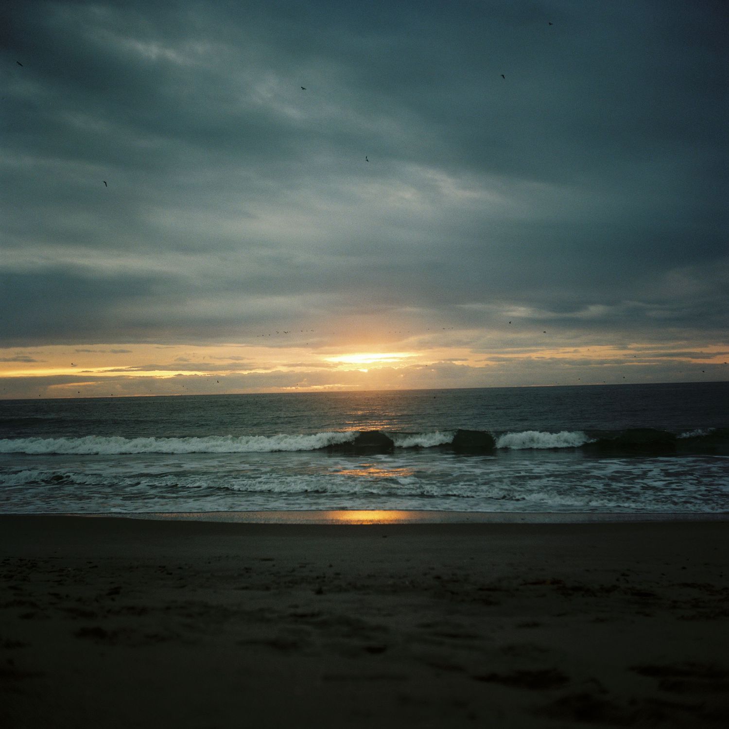 Dramatic ocean sunset with dark clouds and waves crashing against rocks on the horizon.