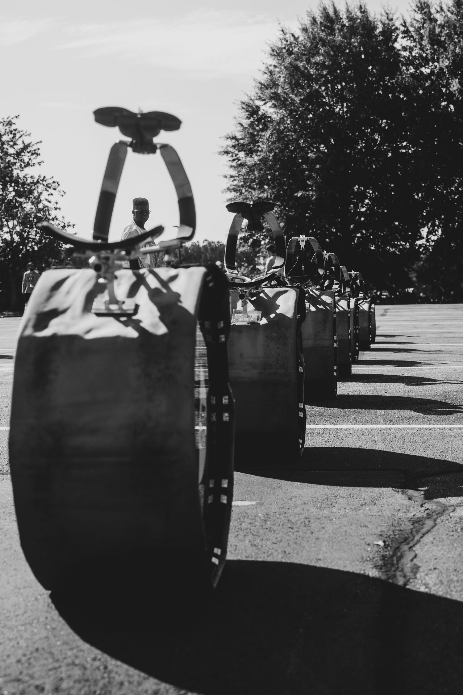 Black and white photo of a row of barrel-shaped vehicles with propellers lined up on a sunny street near trees.