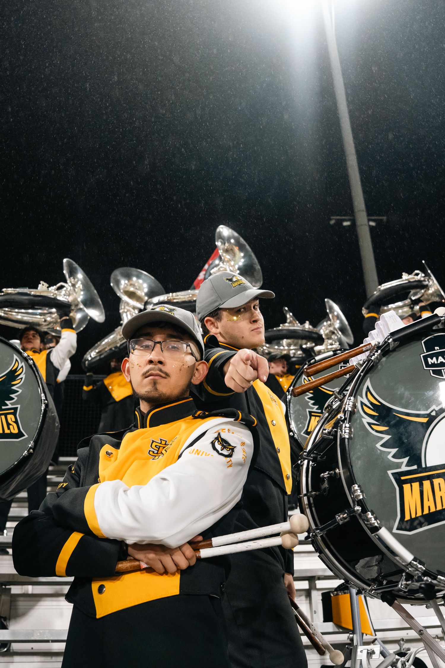 Marching band members in black and gold uniforms perform in the rain at a night football game.