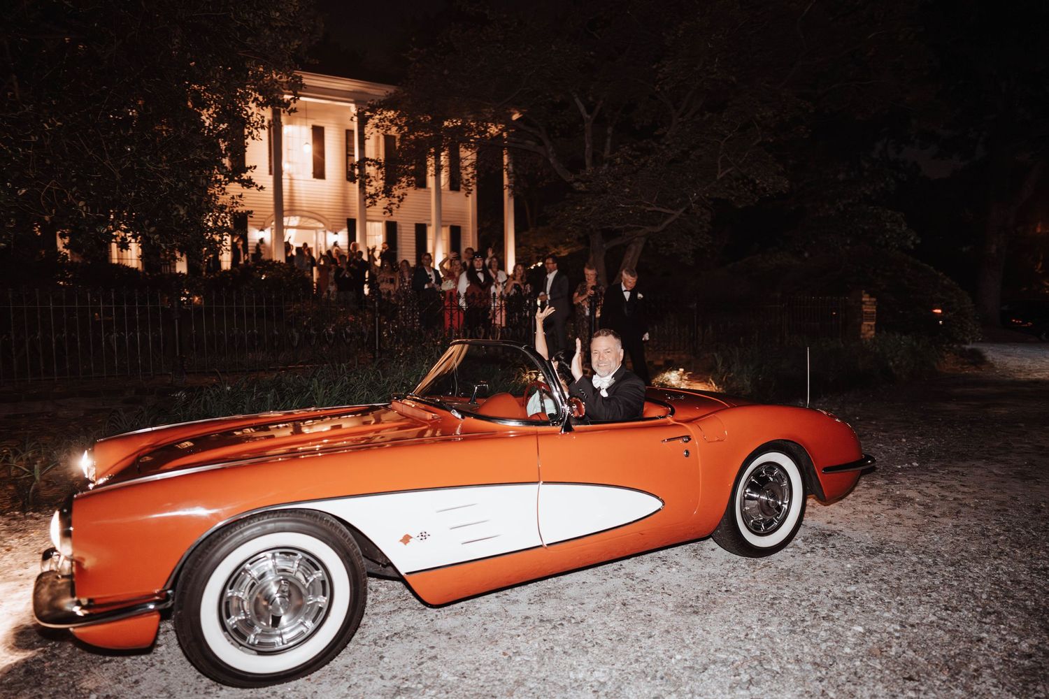 A classic red and white Chevrolet Corvette parked outside an illuminated mansion at night.