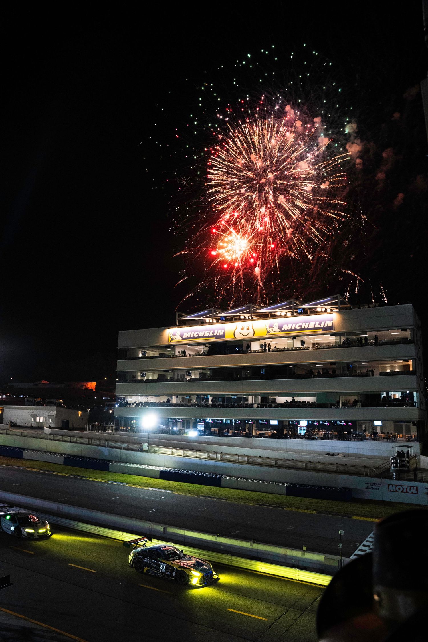 Fireworks explode over illuminated racetrack grandstand during nighttime event.