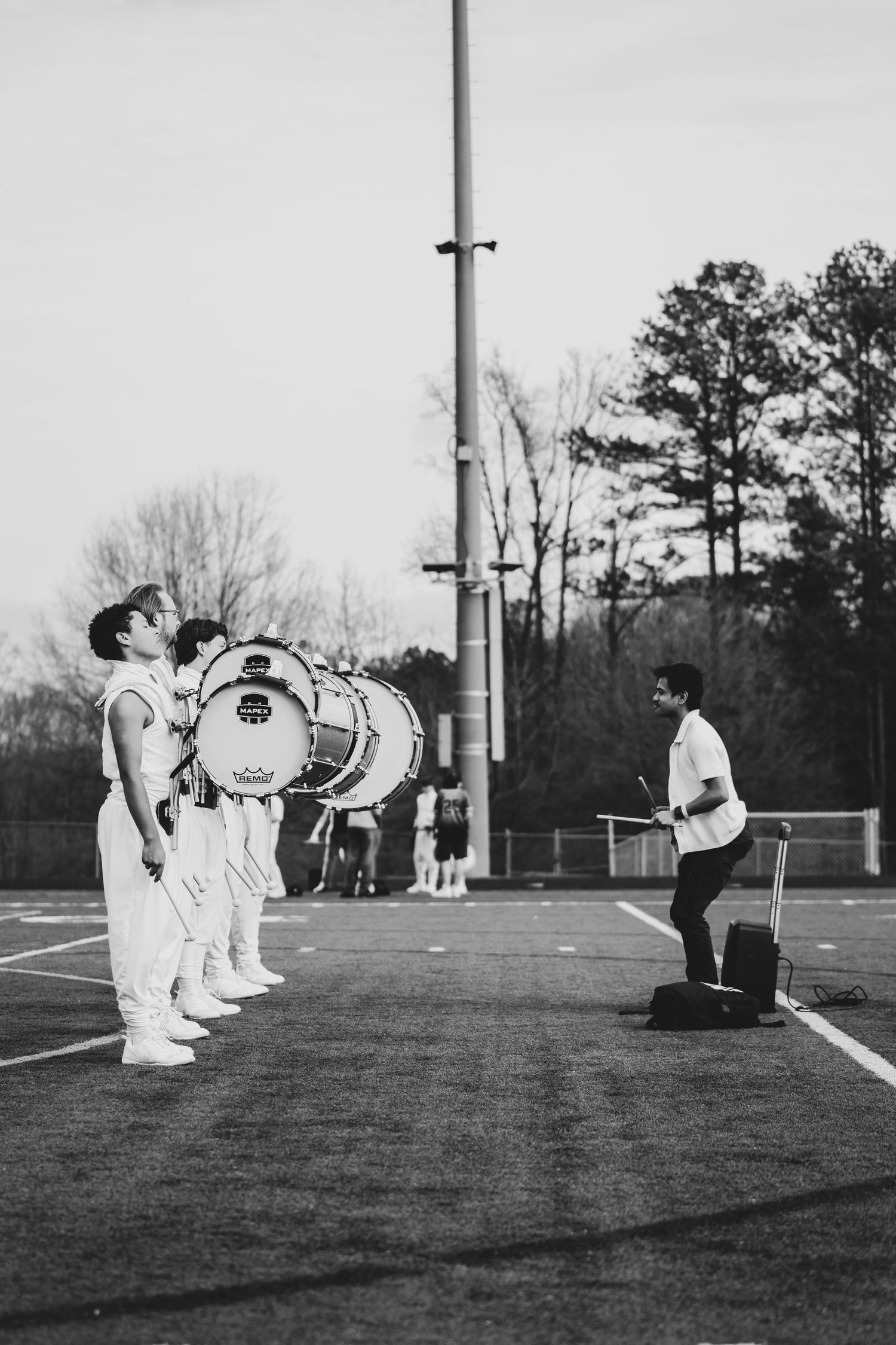 Black and white photo of marching band members with bass drums on a field, facing a conductor during outdoor practice.