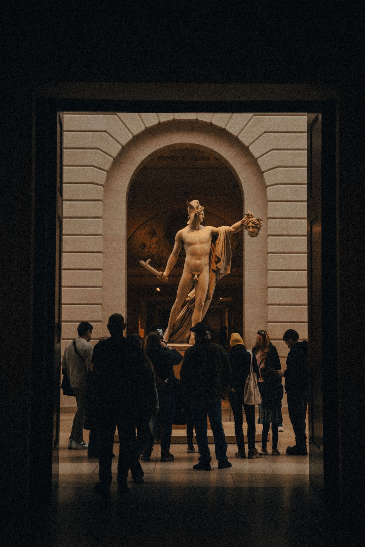 Museum gallery visitors viewing classical sculptures in dramatic lighting.