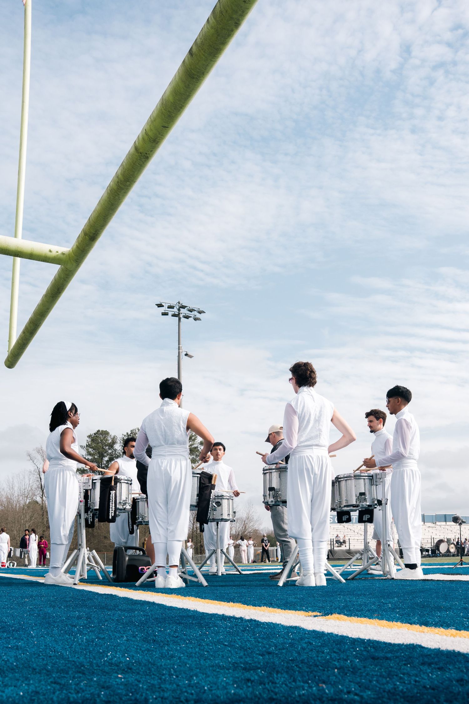 Marching band members in white uniforms perform on a blue turf football field with goal post visible under cloudy sky.