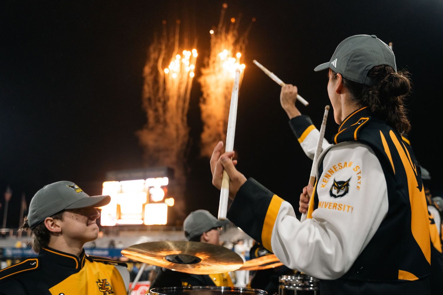 University marching band drummers perform under fireworks at a night football game in black and gold uniforms.