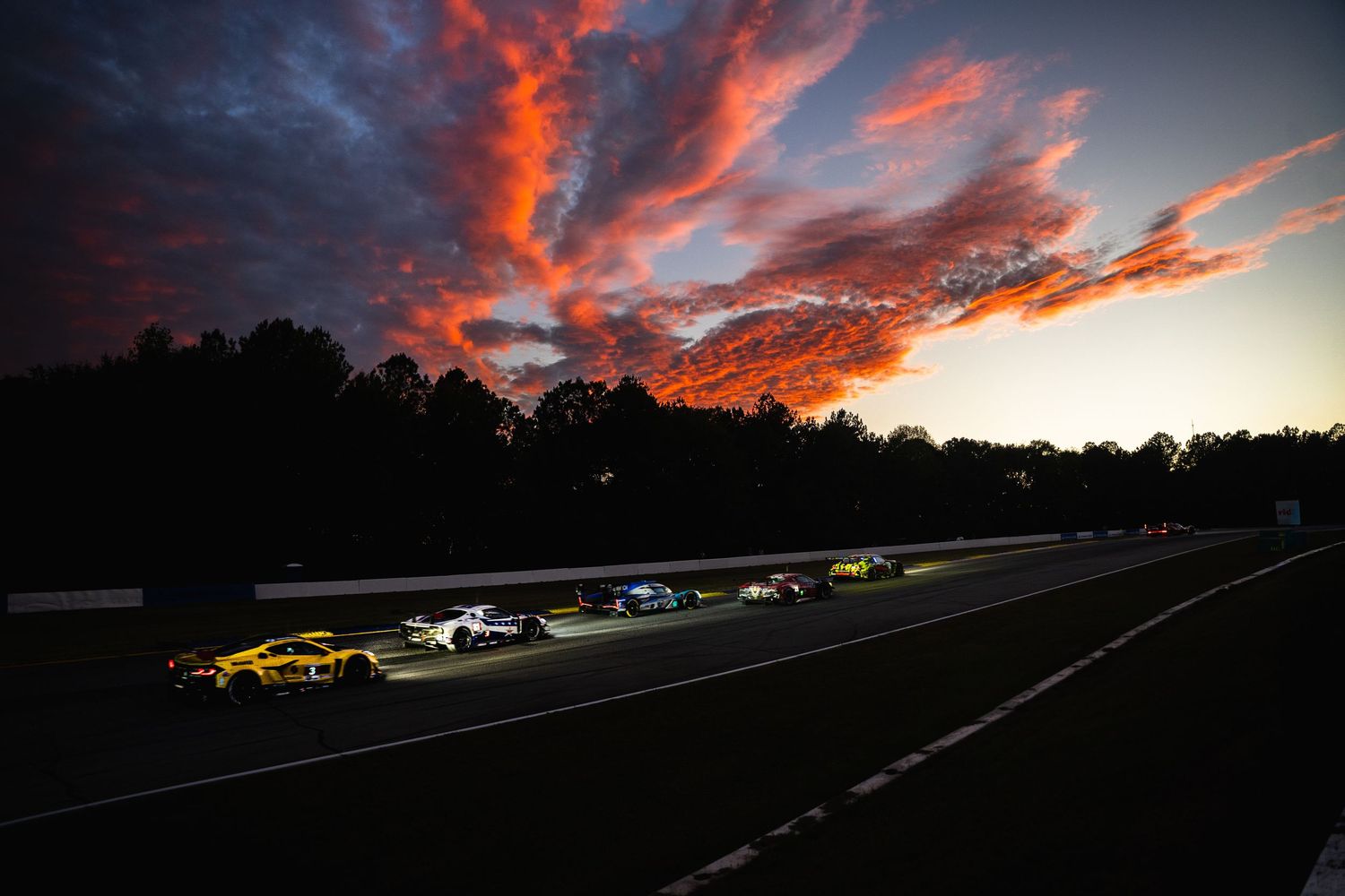 Race cars speed along a track under a dramatic sunset sky with vivid orange and red clouds at dusk.