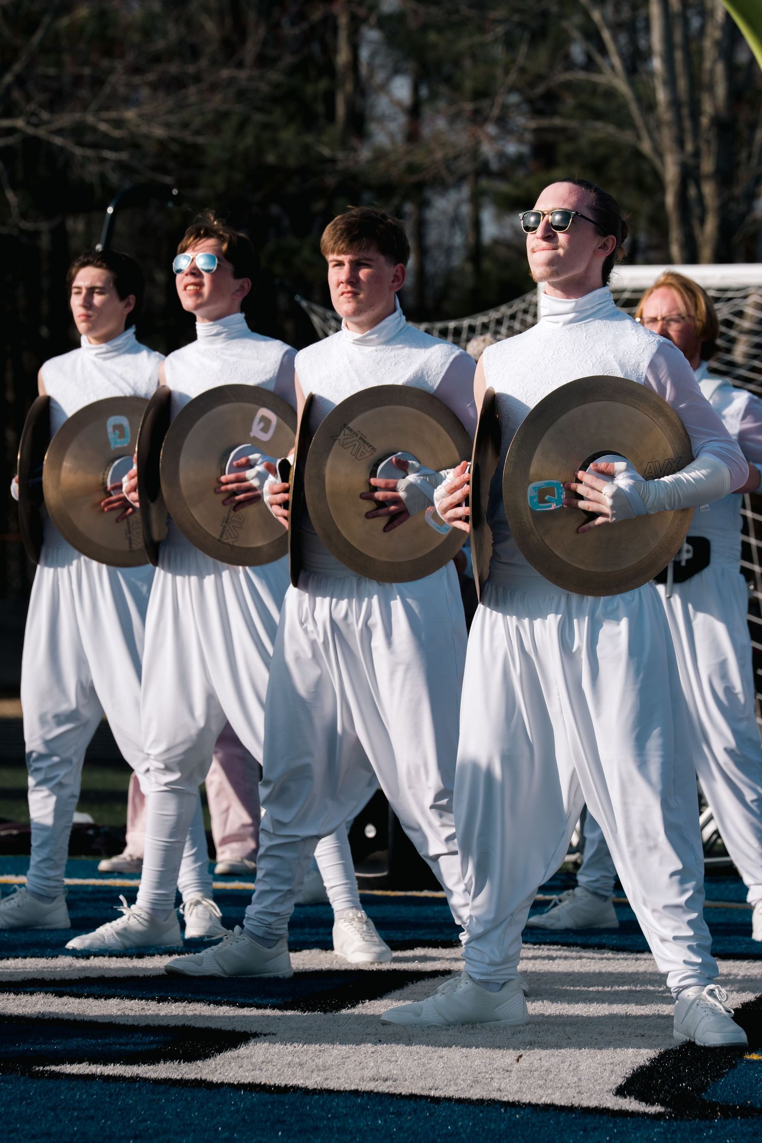 Four performers in white jumpsuits holding cymbals march in formation outdoors during a musical parade performance.