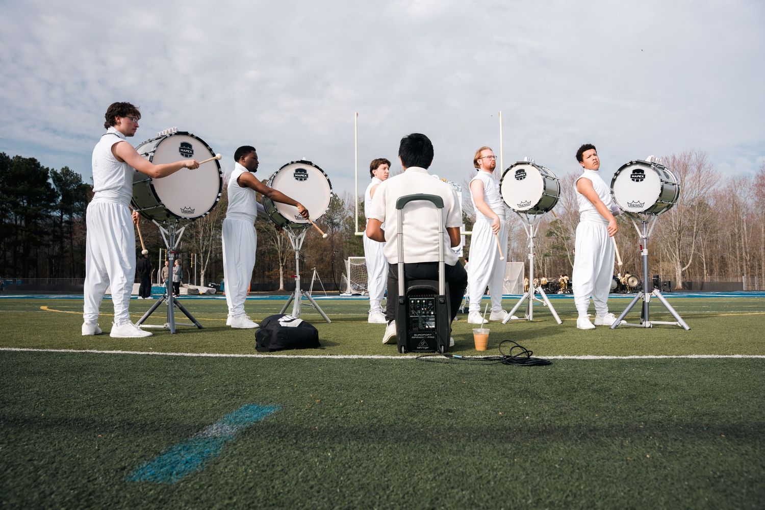 Marching band members in white uniforms playing bass drums on a green turf field outdoors.