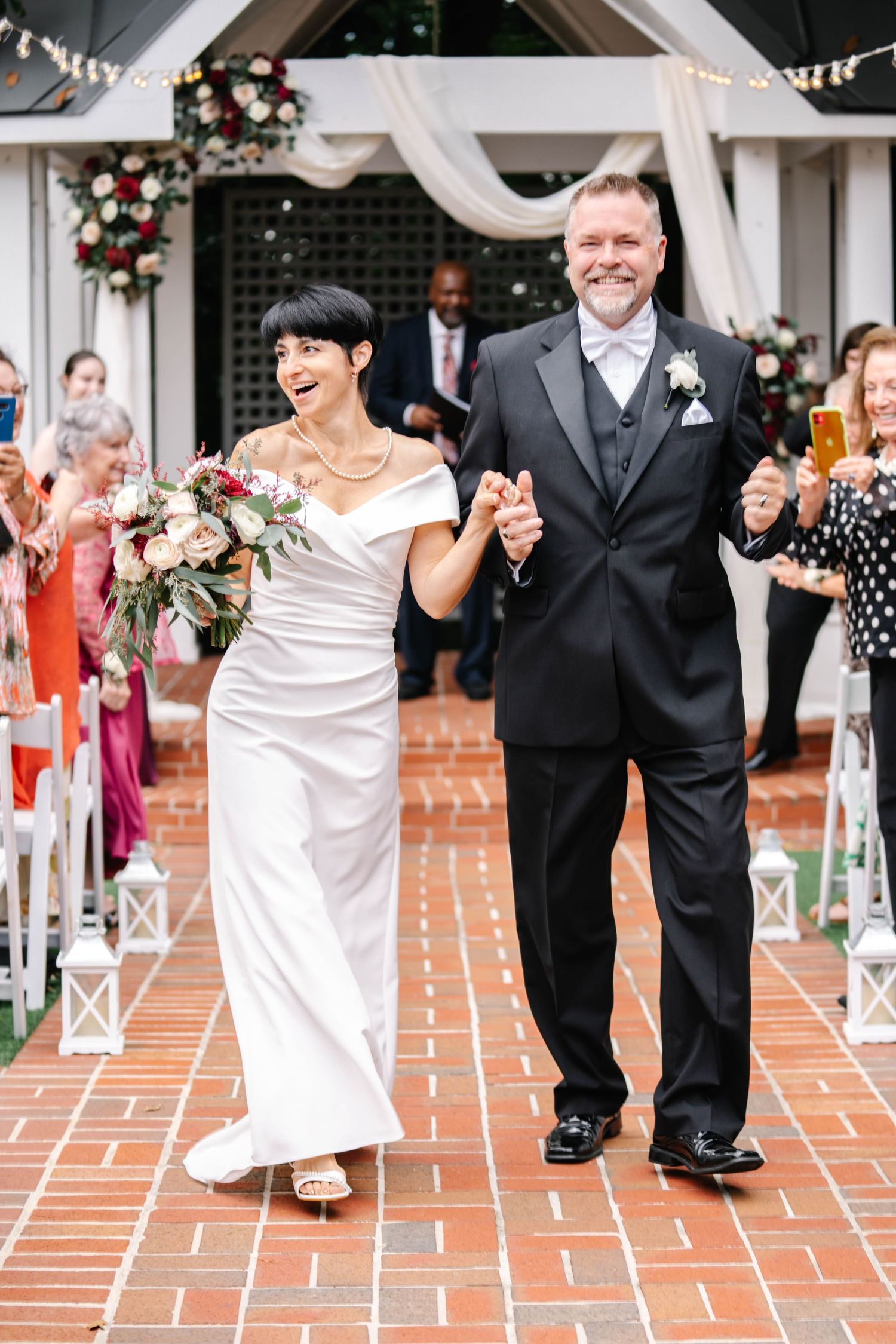 Wedding couple exits ceremony on brick pathway under decorated archway with guests celebrating.