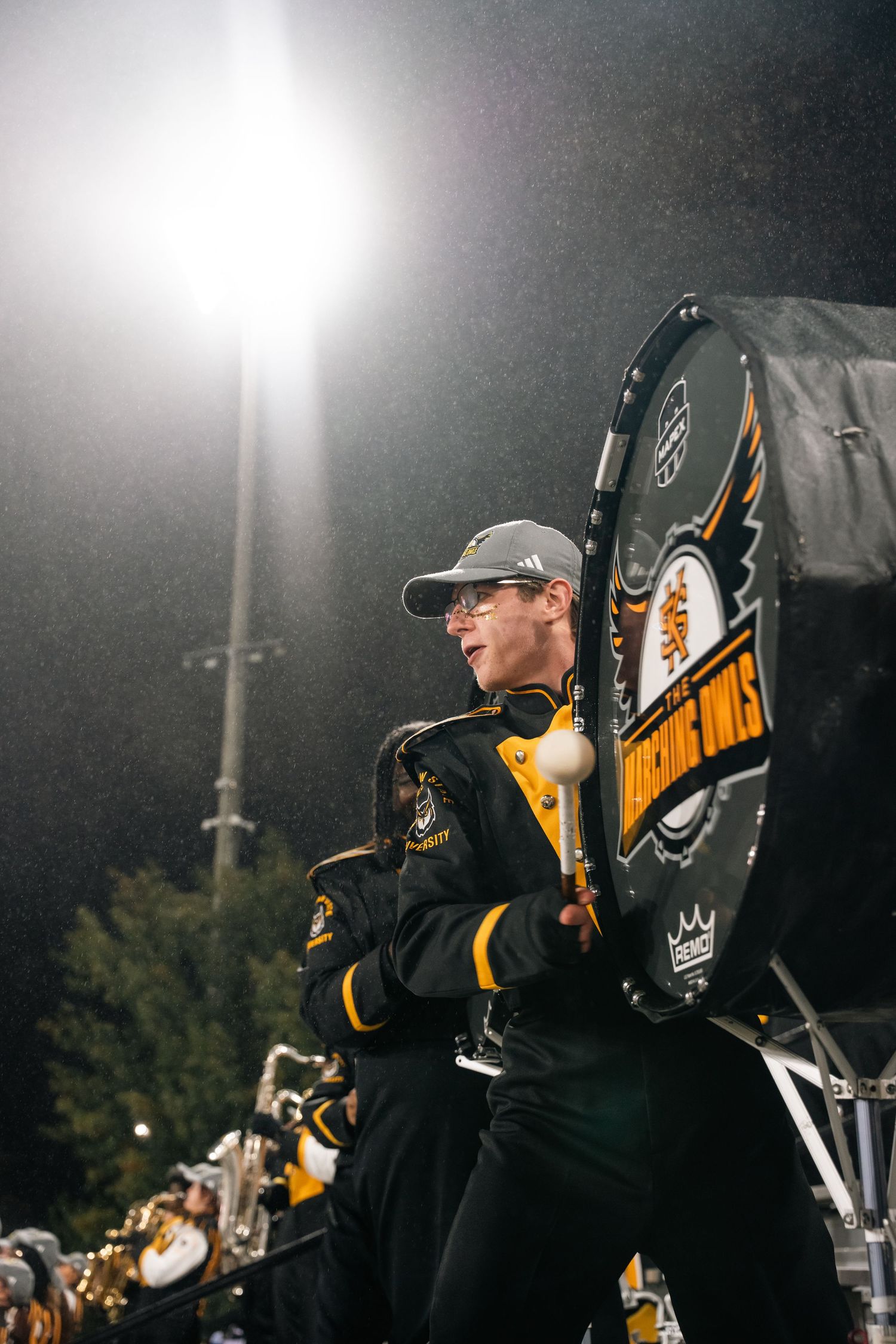 Marching band drummer in black and gold uniform plays bass drum at night game under bright stadium lights.