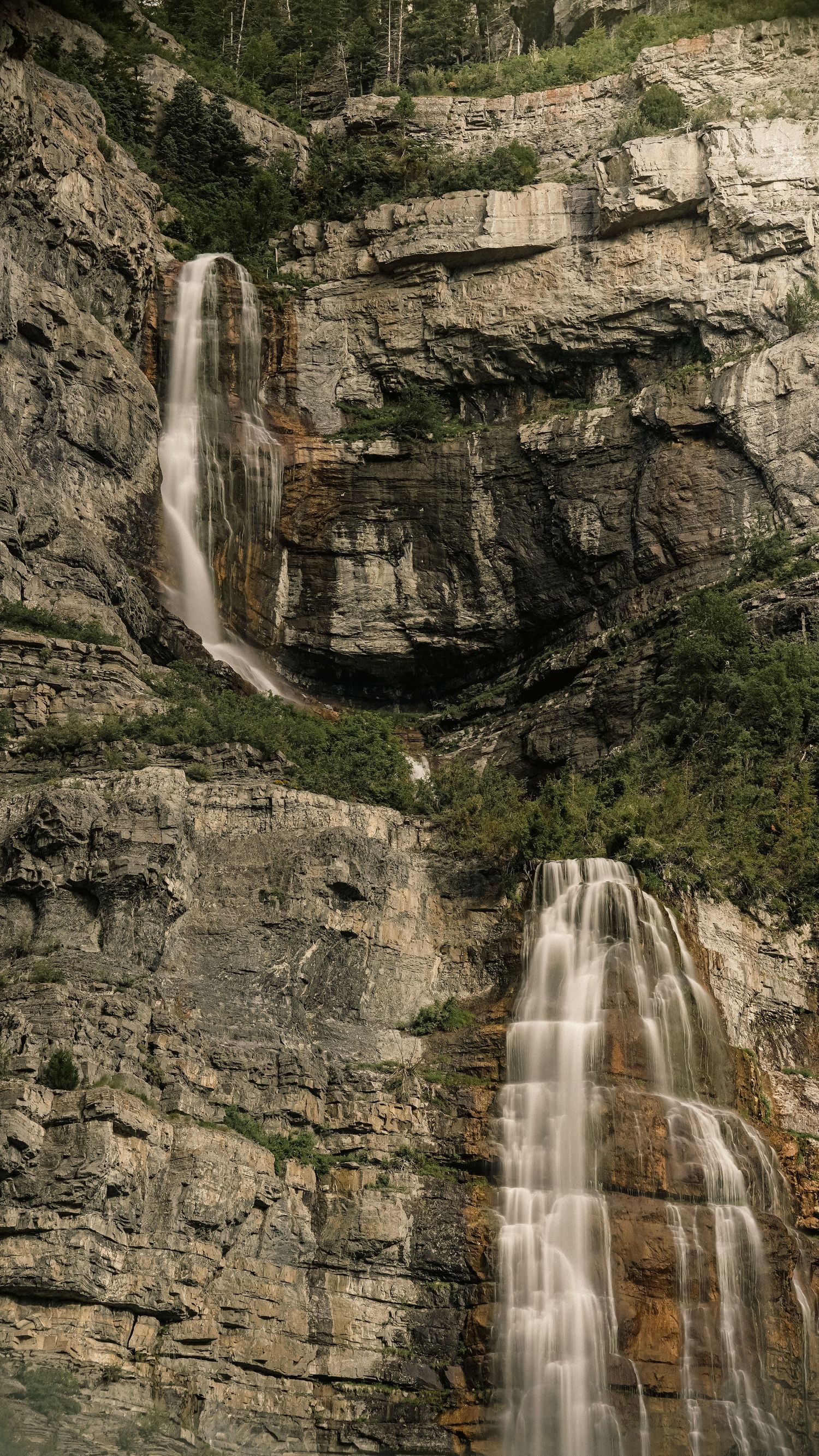 Cascading waterfall flows down massive rocky cliff face surrounded by green vegetation.