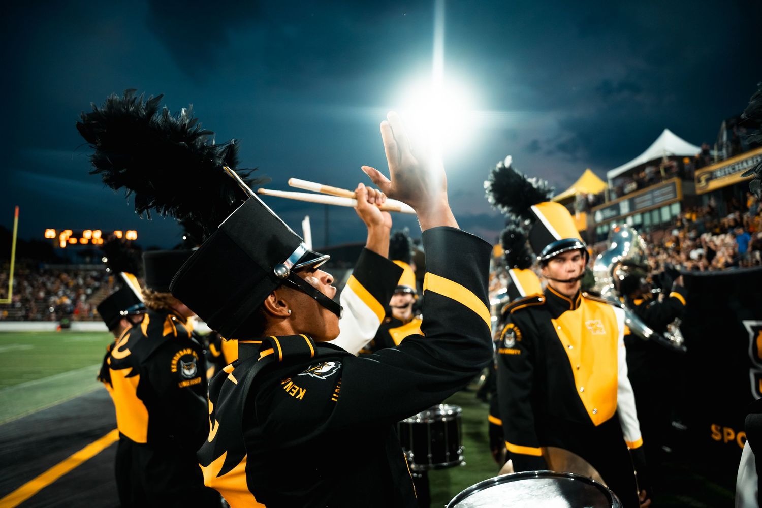 Nighttime performance featuring marching band members with bright stadium lights creating dramatic silhouettes.