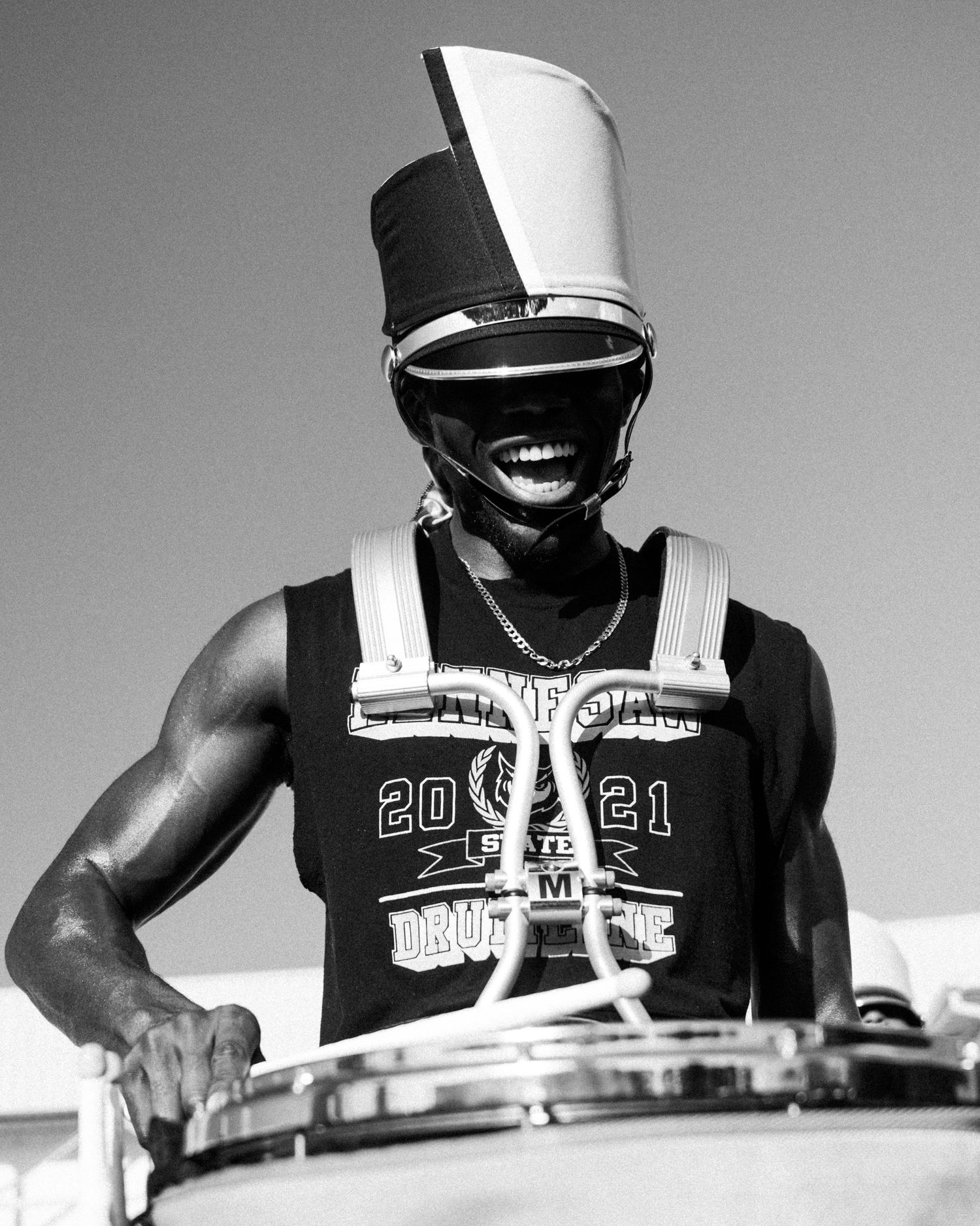 Black and white photo of a smiling drummer in a marching band uniform with tall hat playing snare drum outdoors.