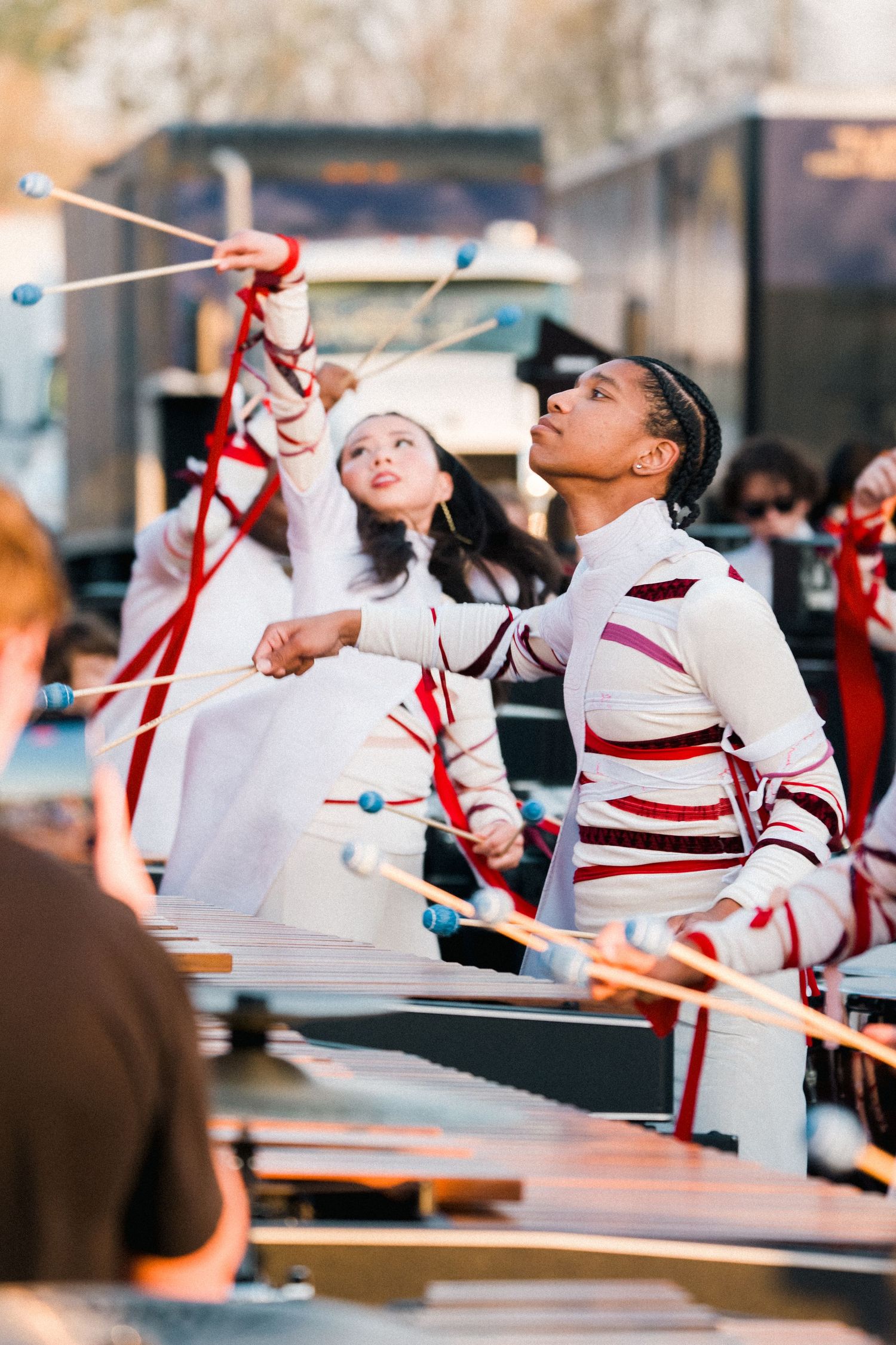 Marching band members in white and red uniforms perform with mallets at an outdoor percussion competition.