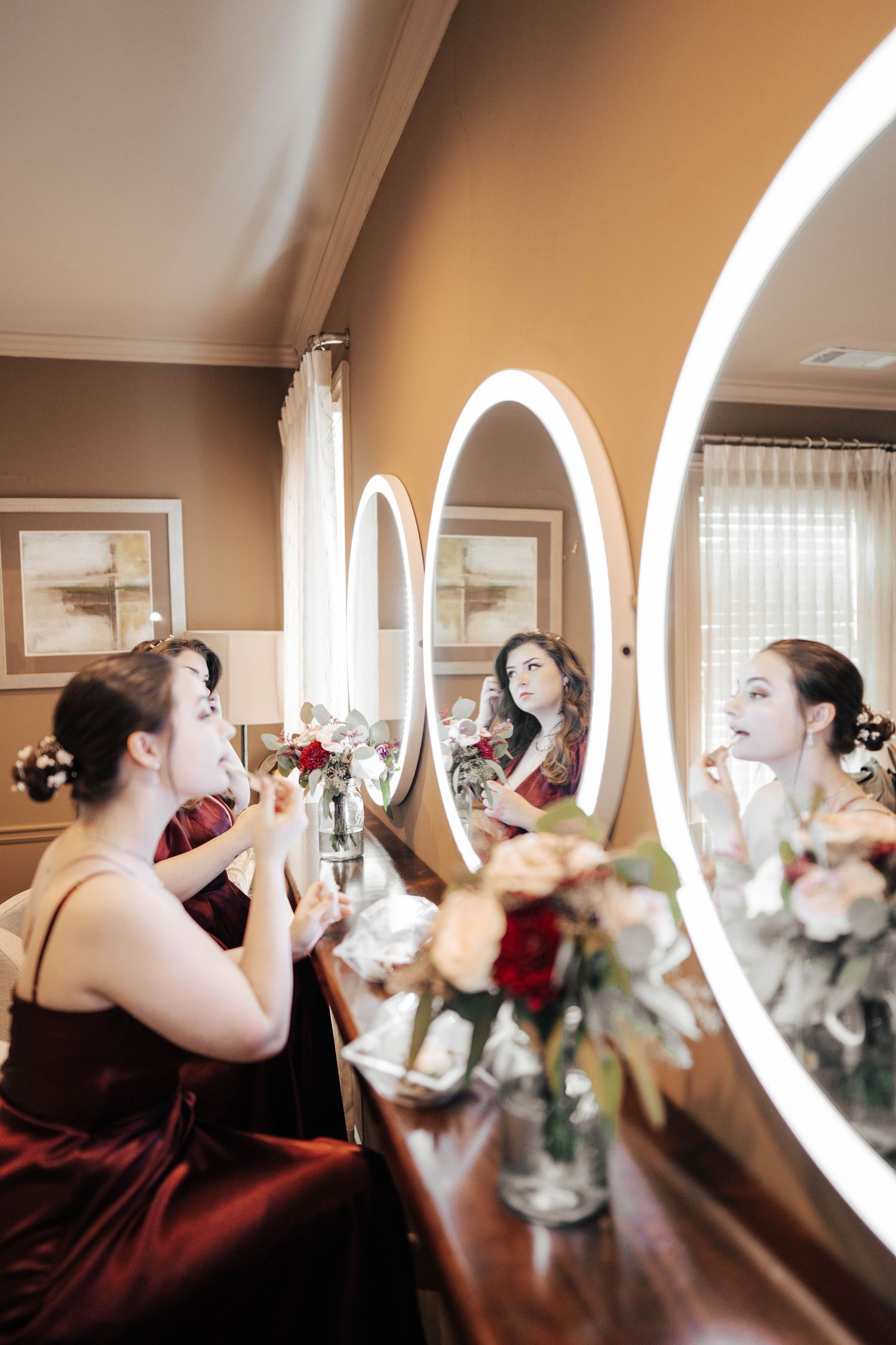 Bridesmaids in burgundy dresses toast with champagne glasses in front of curved mirror.