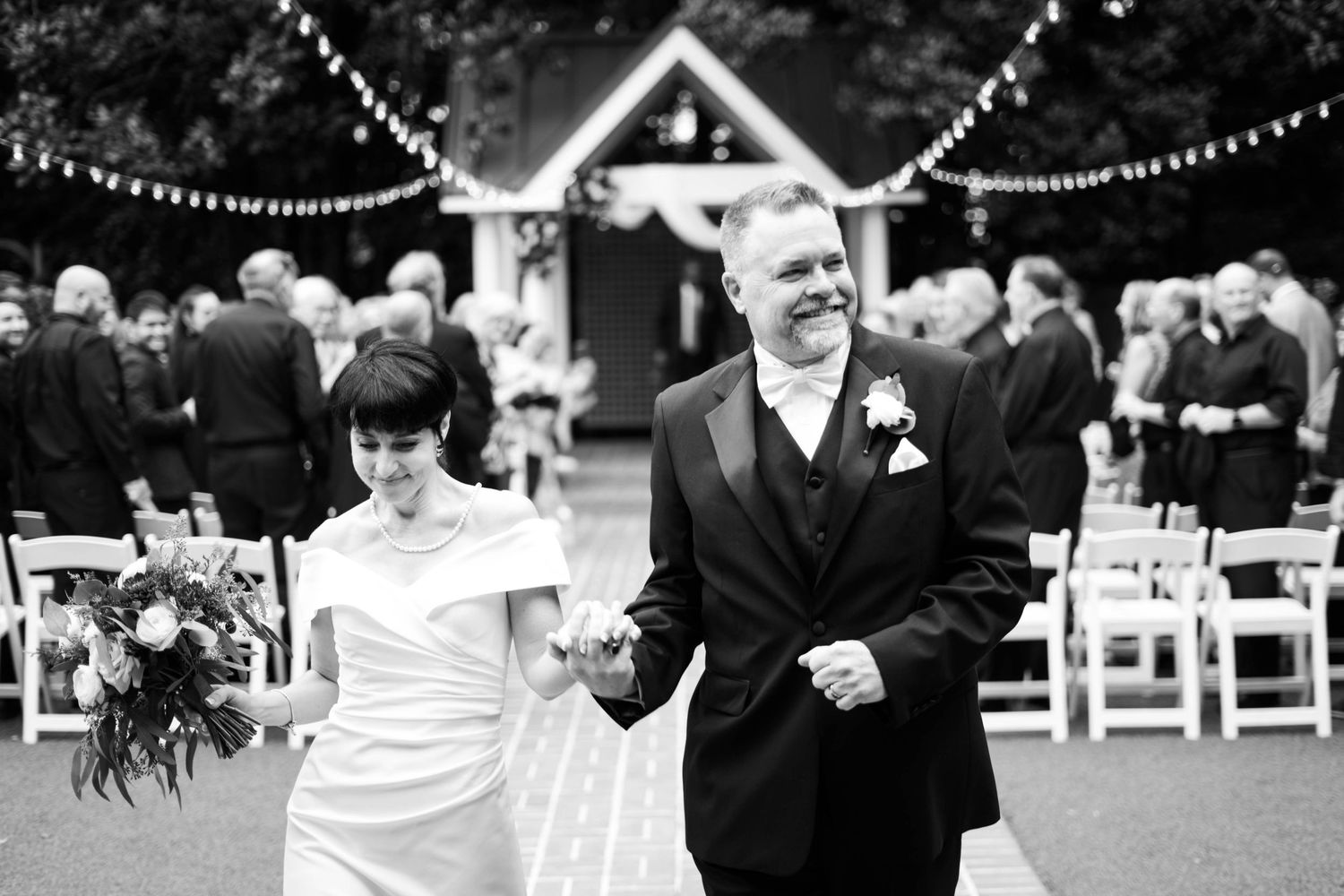 A joyful recessional walk down an outdoor aisle with string lights and white chairs at a garden wedding ceremony.
