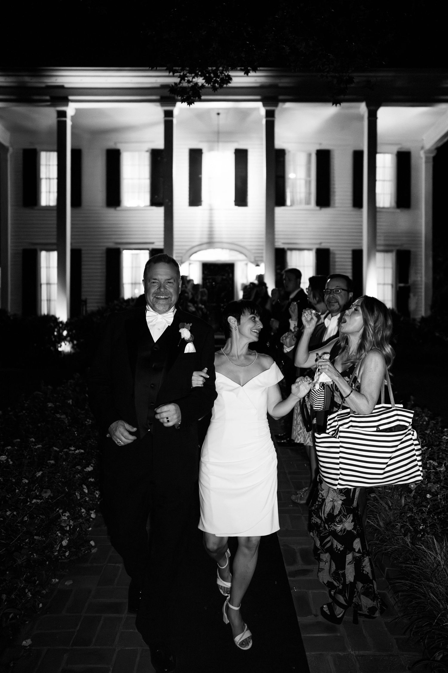 A nighttime celebration outside a grand white columned mansion with party guests.