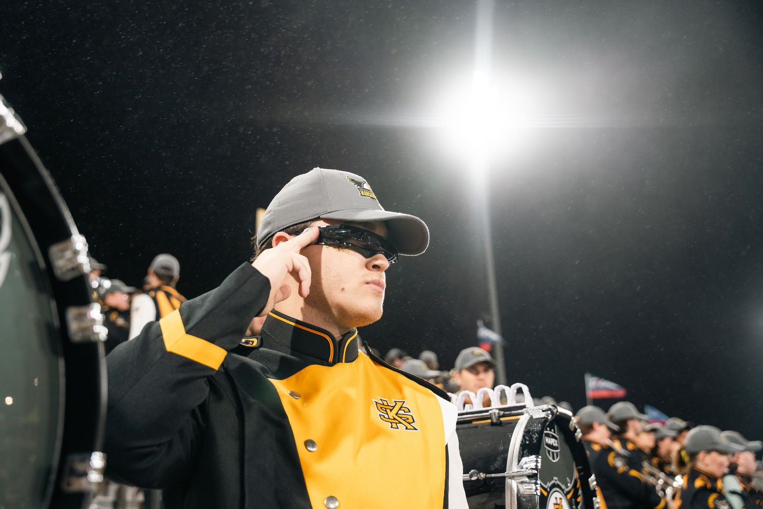 Band performers in yellow uniforms raise their arms in synchronized motion under stadium lights.