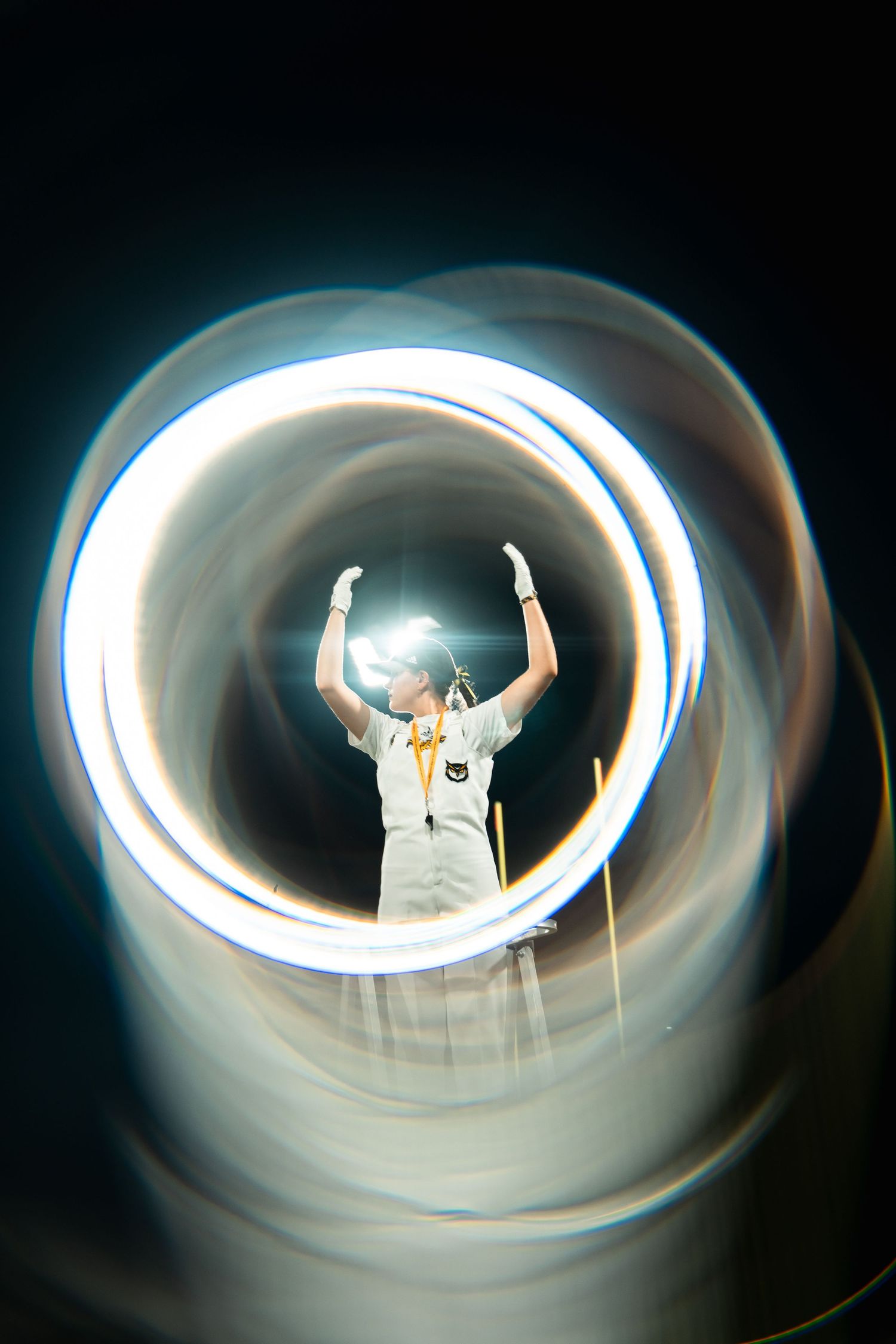 Person in white uniform with raised arms framed by glowing circular light trails against a dark background.