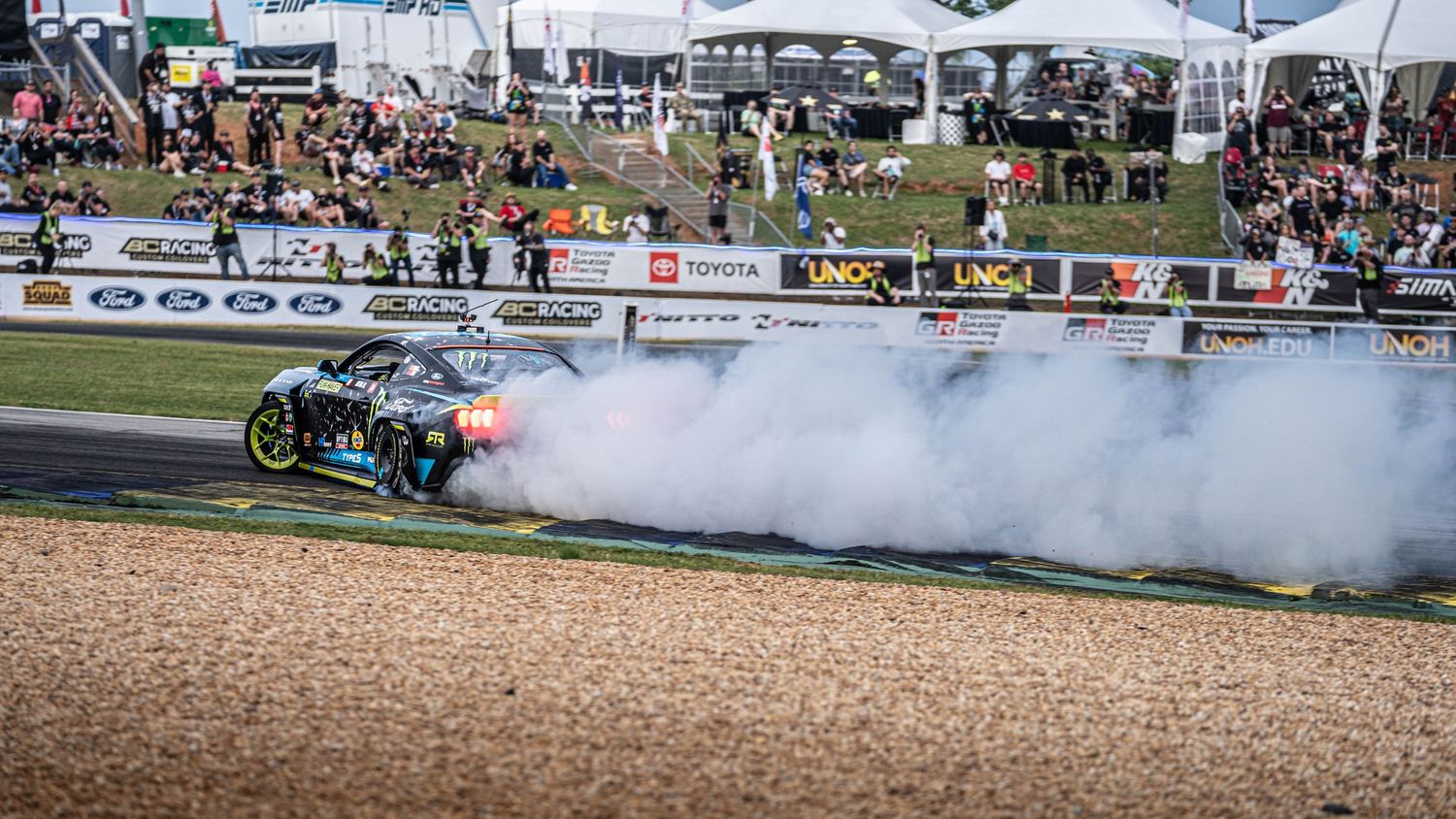 Panoramic view of motorsport event with crowd and tire smoke on race track.