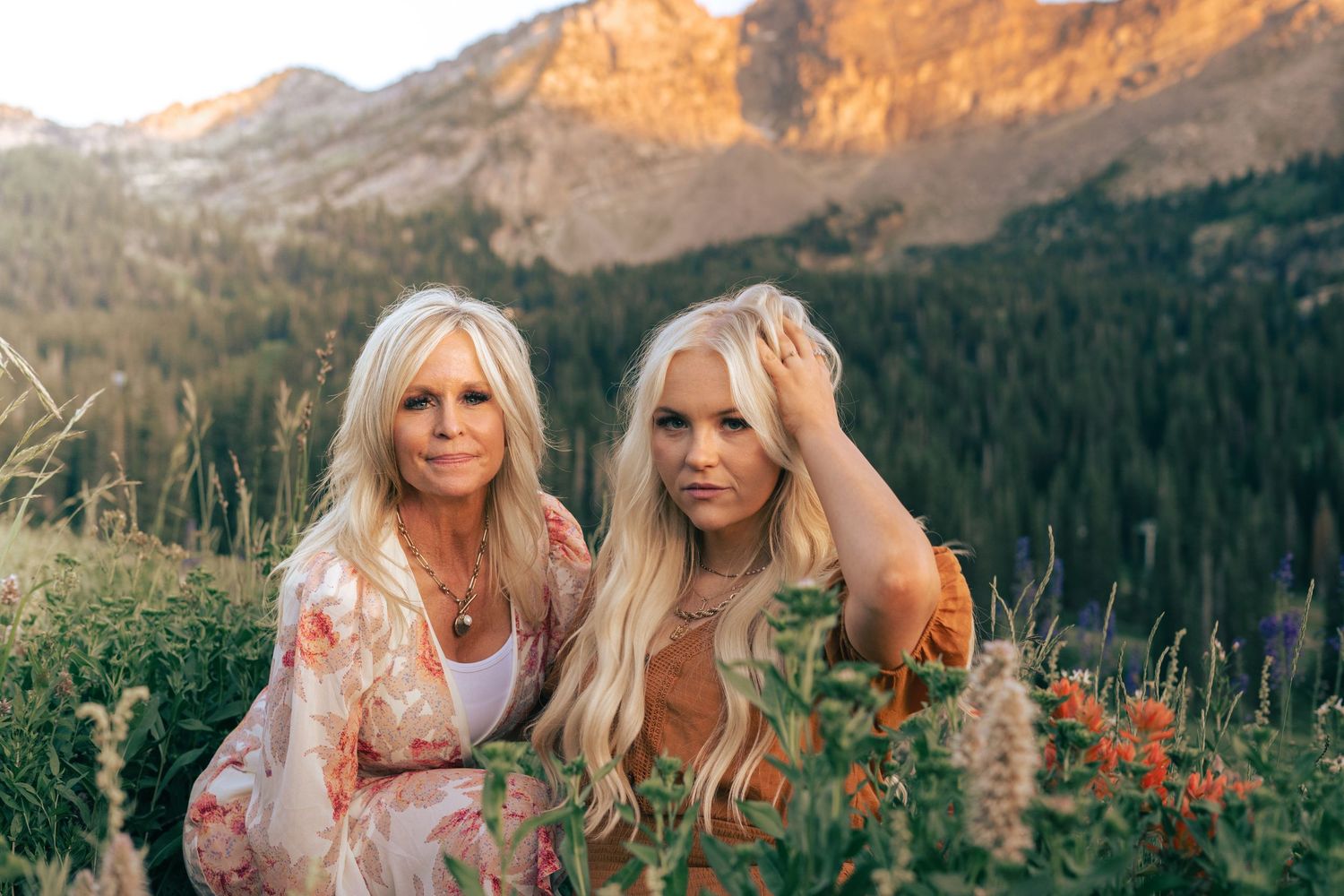 Couple in a floral dress and casual attire pose together against a scenic mountain backdrop at sunset.