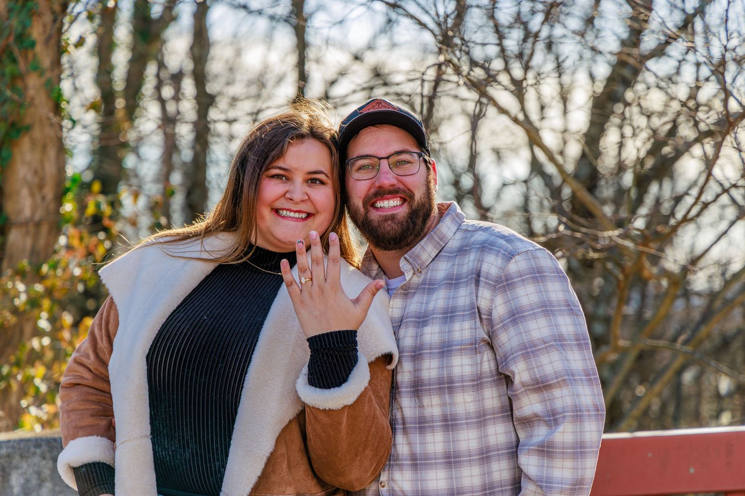Happy couple smiling outdoors showing off engagement ring, surrounded by autumn trees in a park setting.
