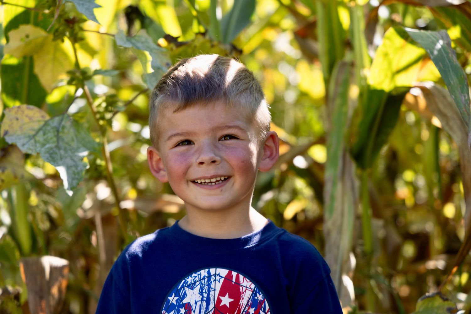 A smiling young boy with short hair wearing a patriotic navy blue t-shirt, standing outdoors near green corn plants.