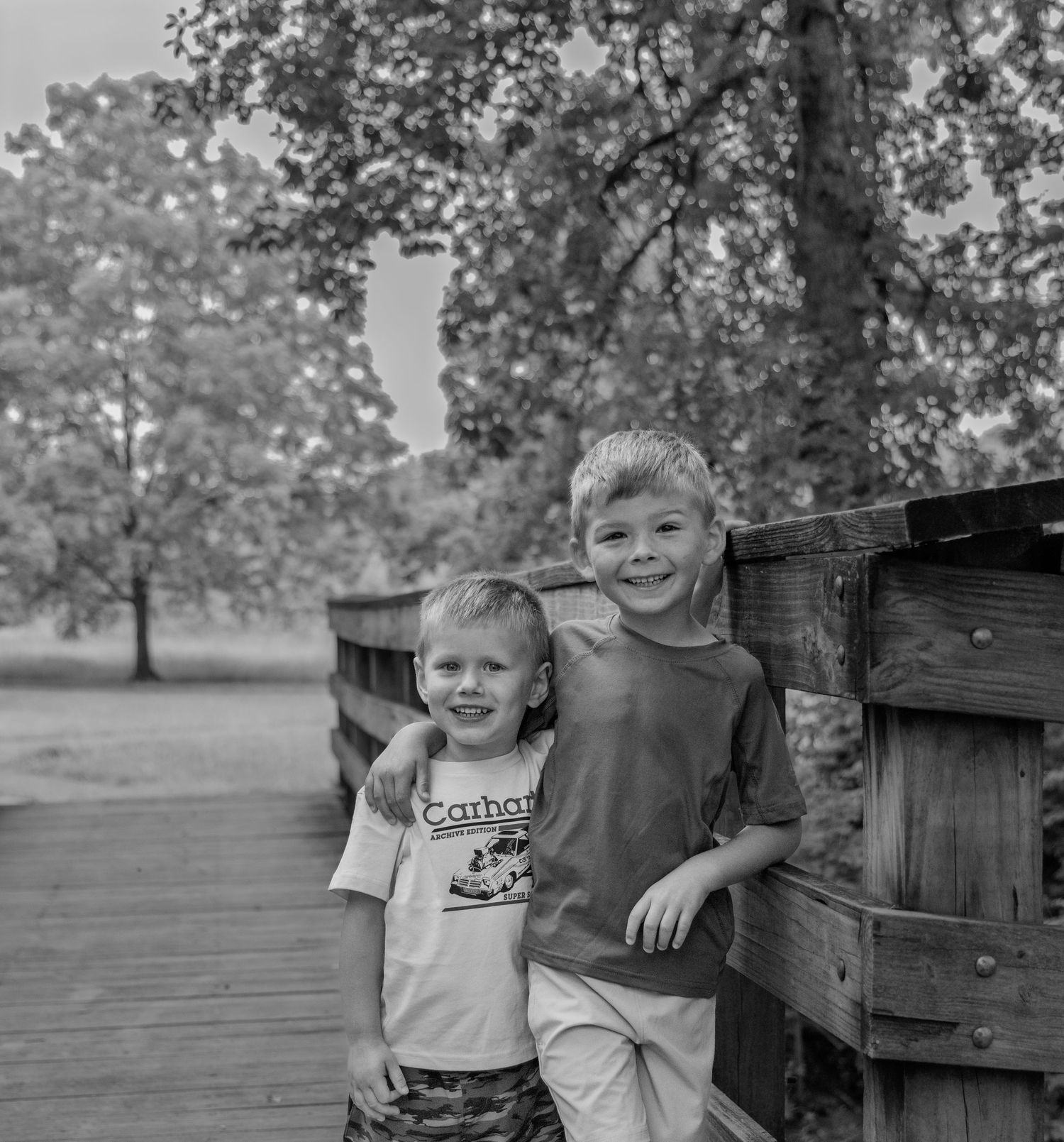 Two young boys smiling on a wooden bridge surrounded by trees in a black and white outdoor photo.
