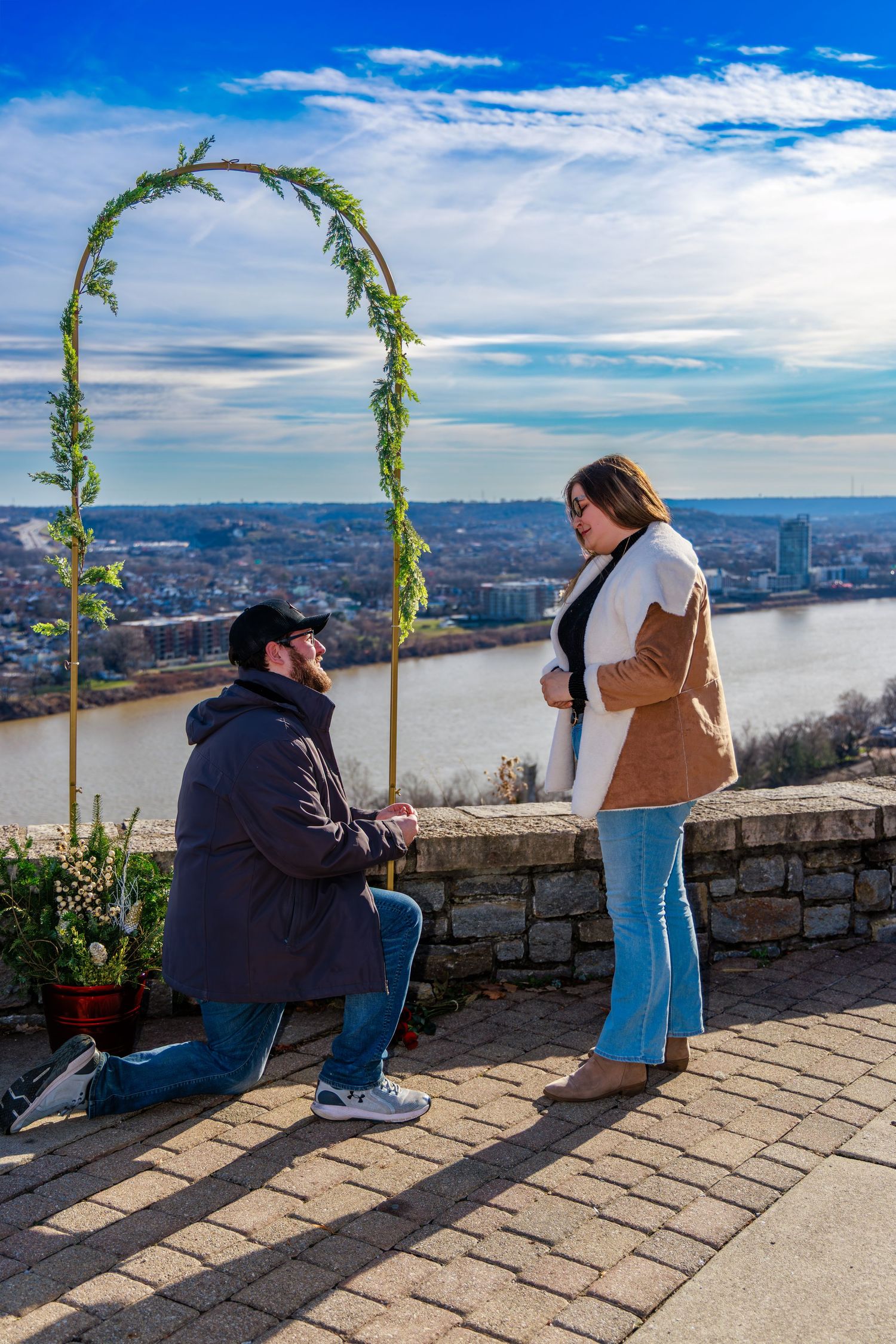 Man proposes on one knee to woman outdoors near river overlook with decorative arch and city skyline backdrop.