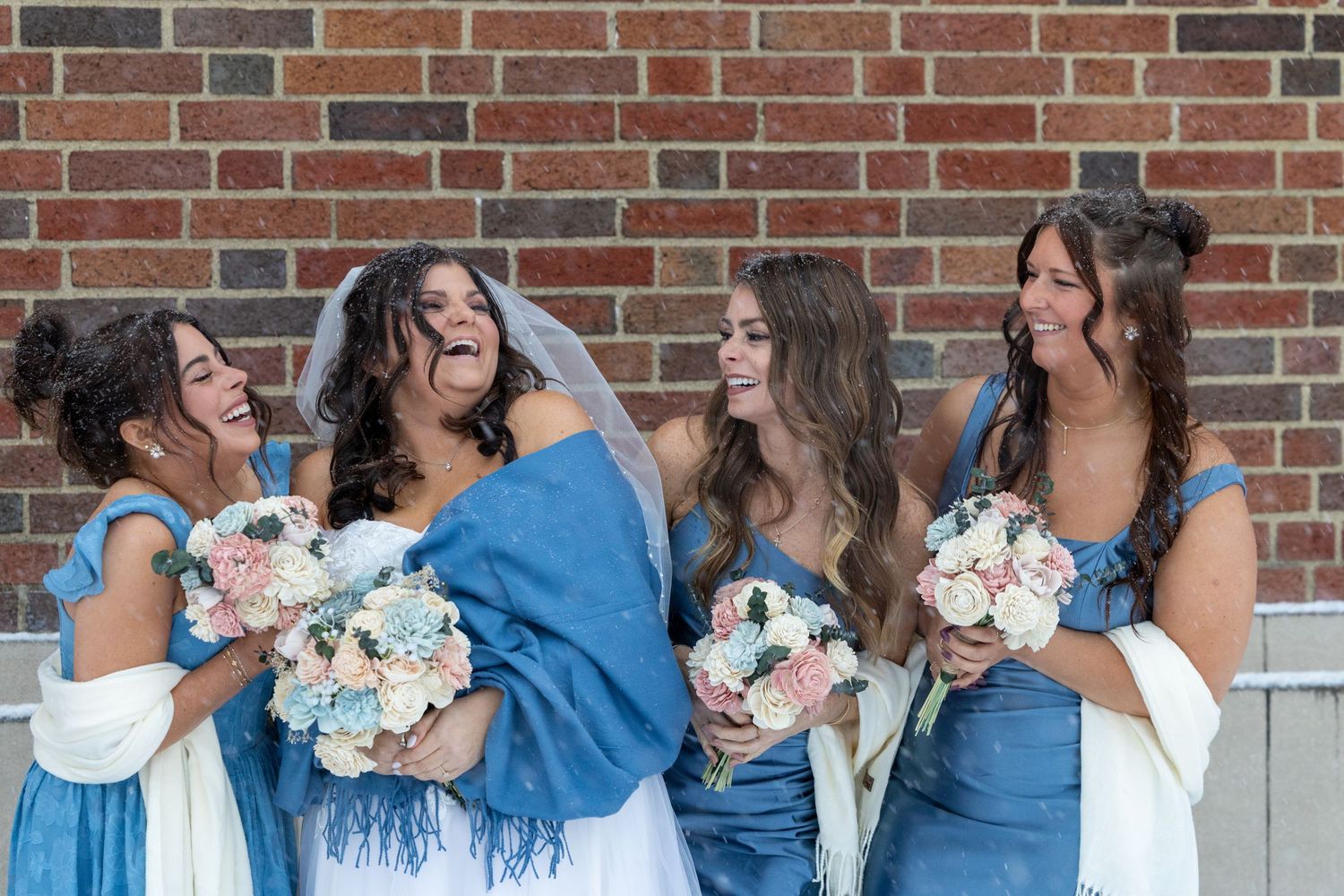 Bride laughing with three bridesmaids in blue dresses holding floral bouquets against a brick wall.