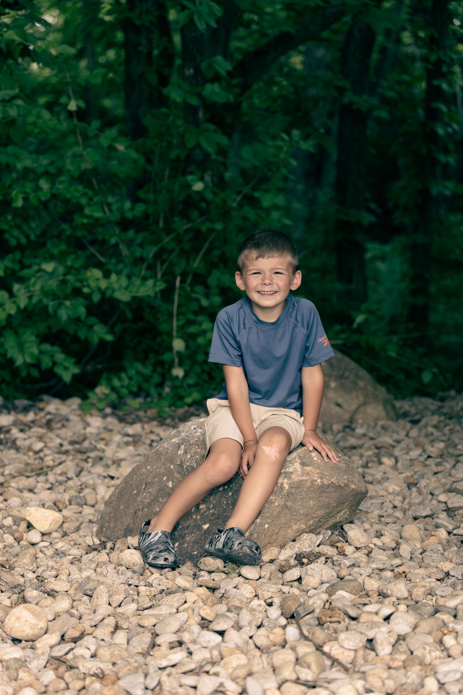Smiling young boy sitting on a large rock surrounded by gravel and lush green trees in a natural outdoor setting.
