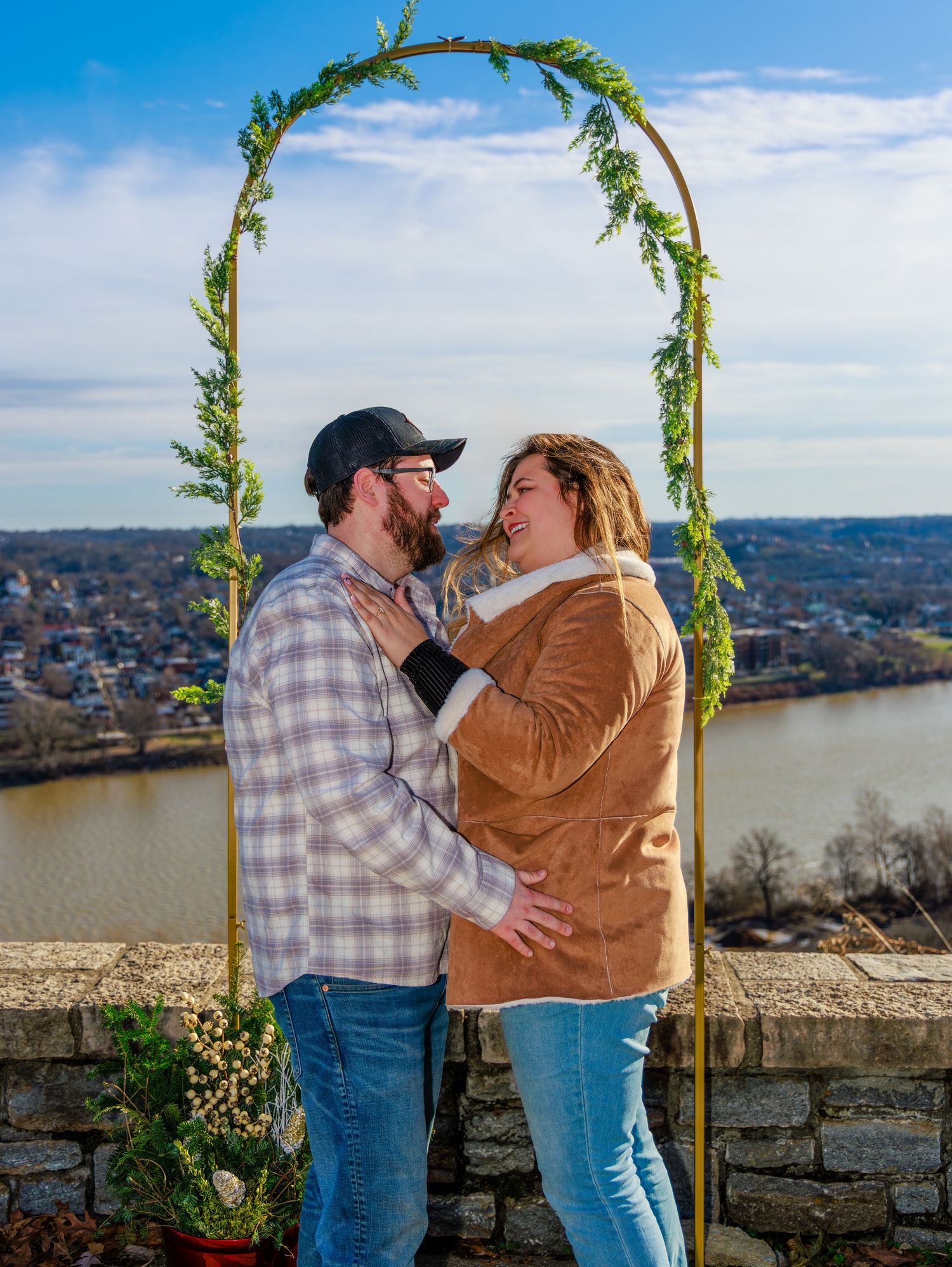 Couple embracing under a floral arch with a river and town view behind them on a sunny day.