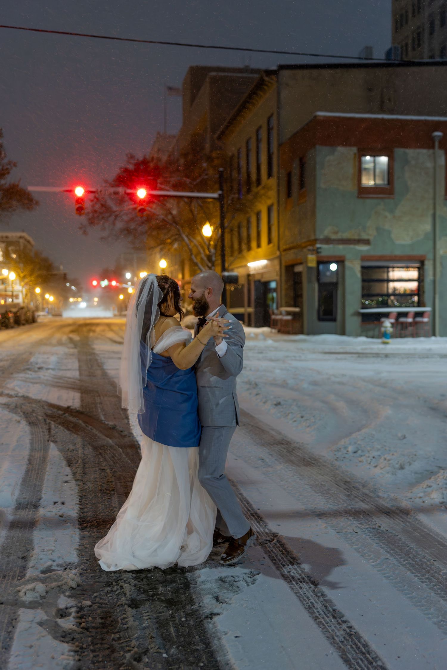 Bride and groom share a romantic dance on a snowy city street at night, surrounded by glowing traffic lights.