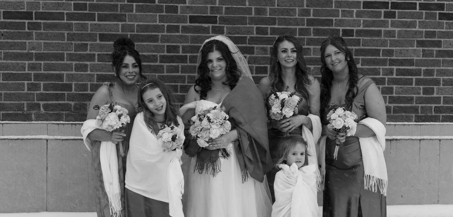 Black and white wedding photo of bride with bridesmaids and flower girls holding bouquets in front of brick wall.