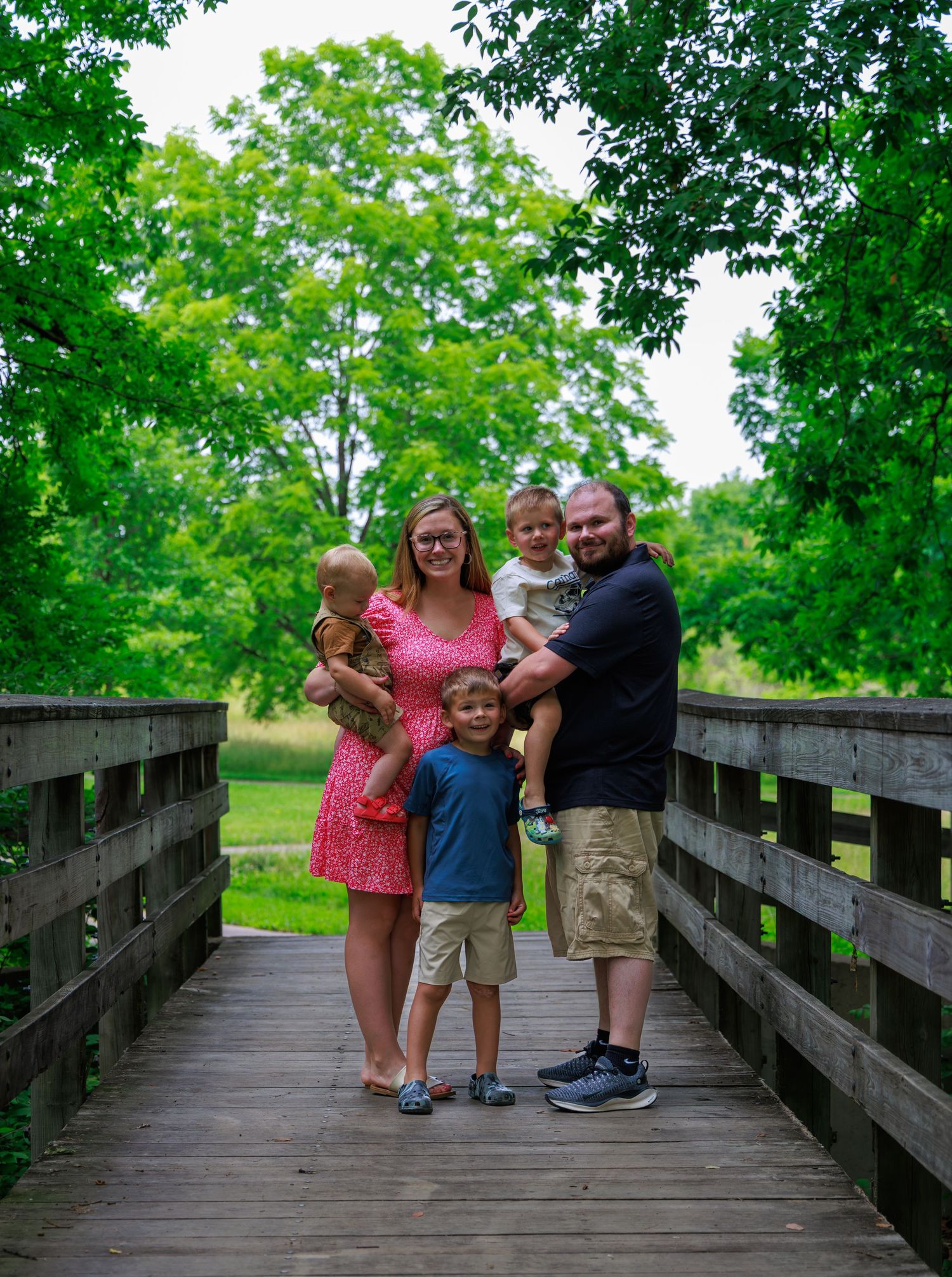 Family of five posing on a wooden bridge surrounded by lush green trees on a sunny day.