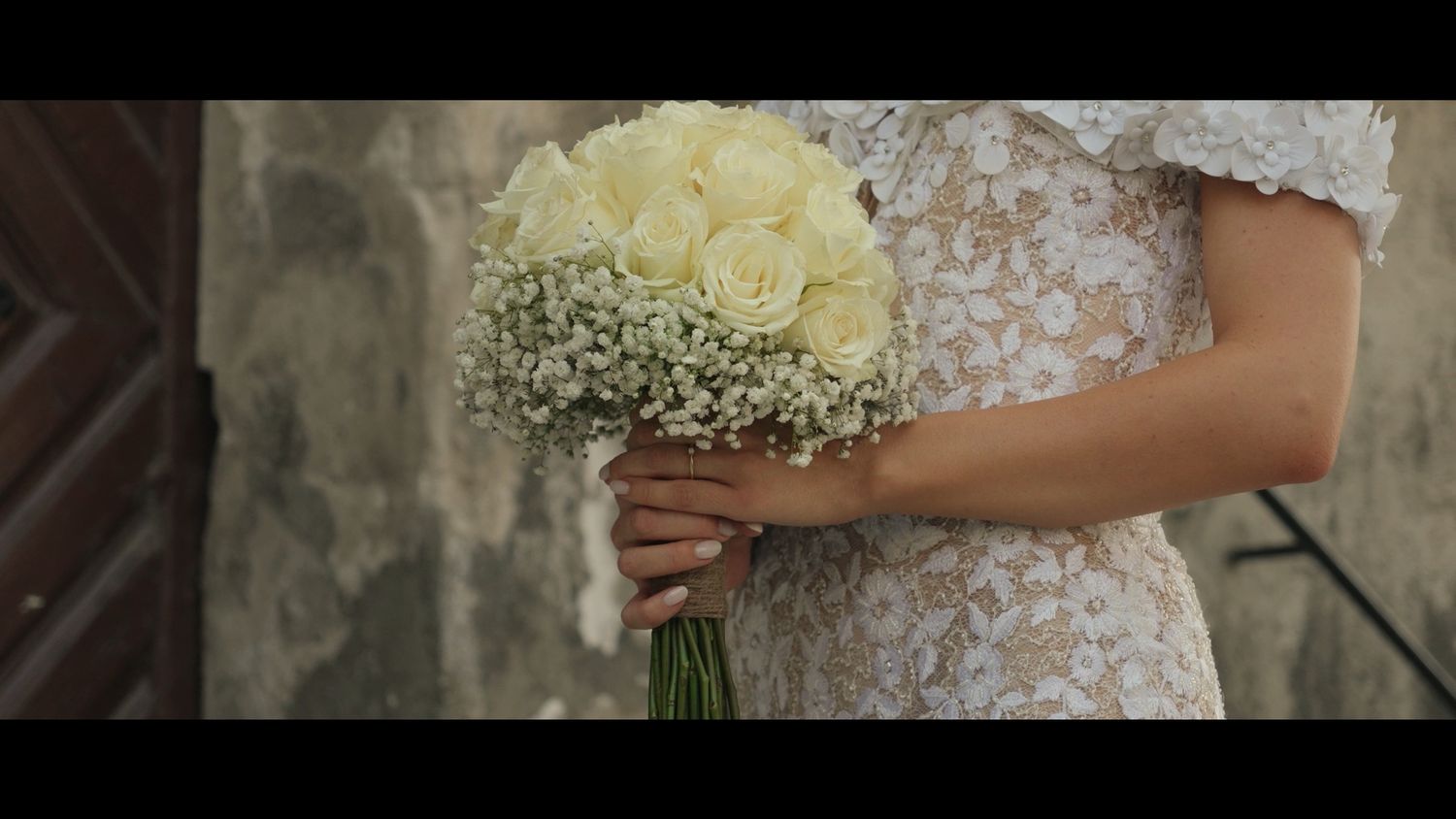 Bride in lace gown holds white floral bouquet during outdoor wedding ceremony.