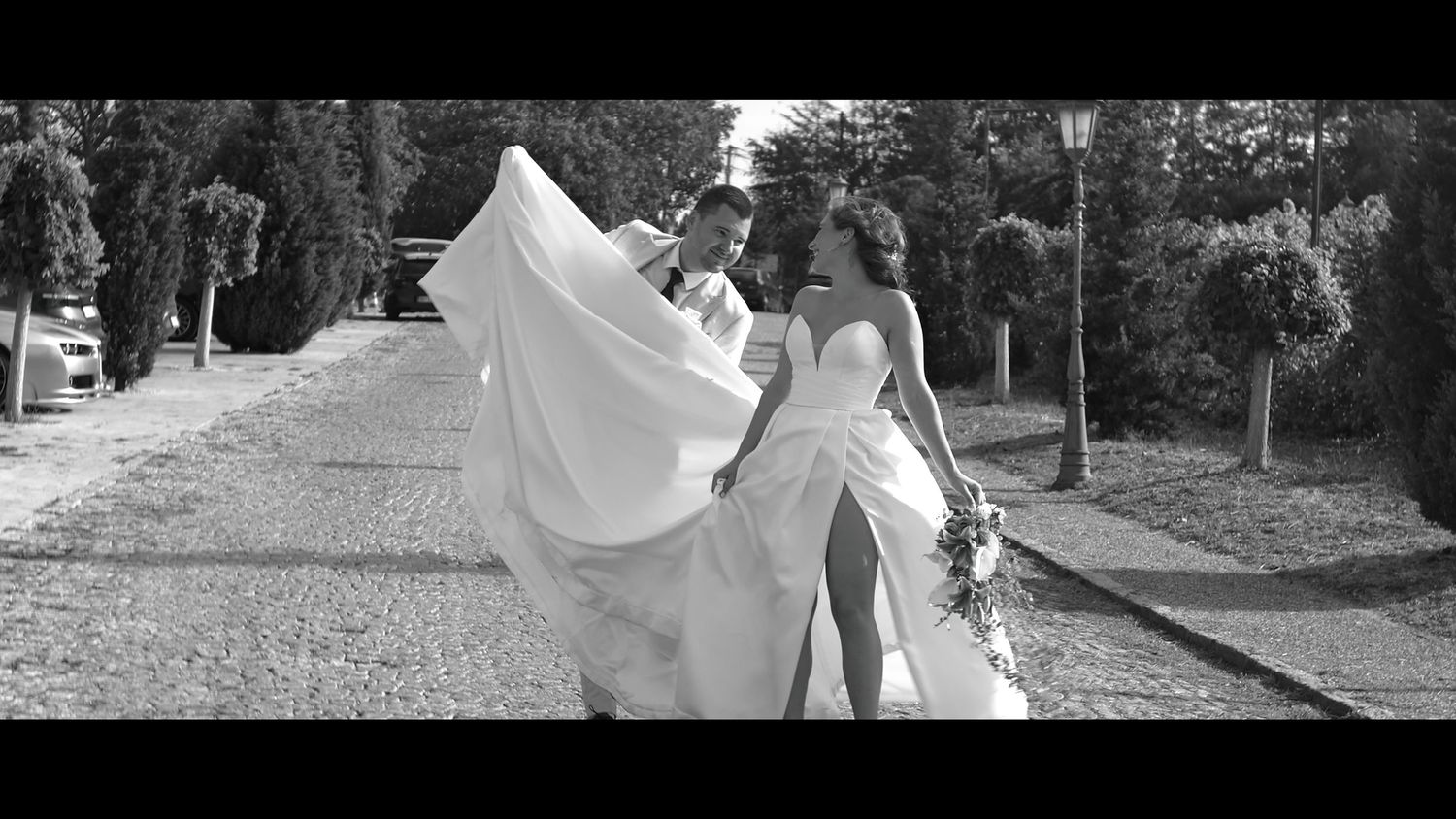 A flowing white wedding dress billows in the breeze on a tree-lined path.