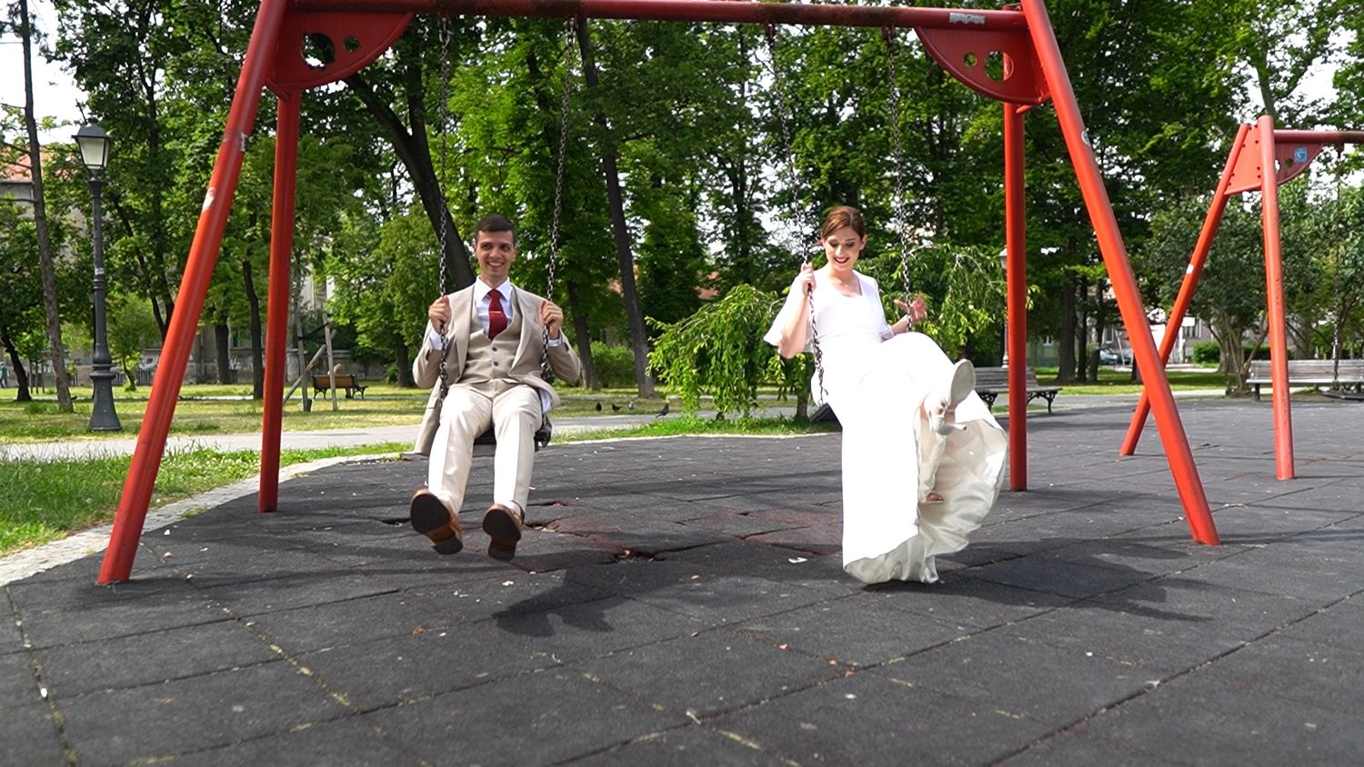 Wedding couple posing playfully on red playground swings in a park.