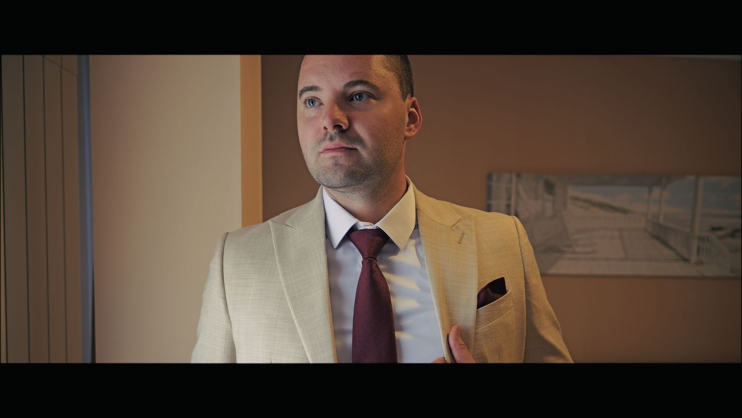 Person wearing beige suit with burgundy tie standing in room.