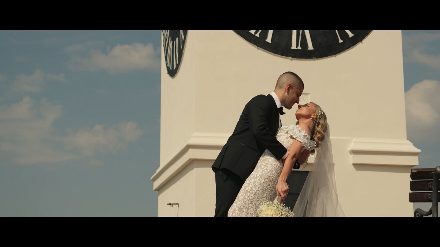 Wedding couple embracing against dramatic sky with vintage clock in background.