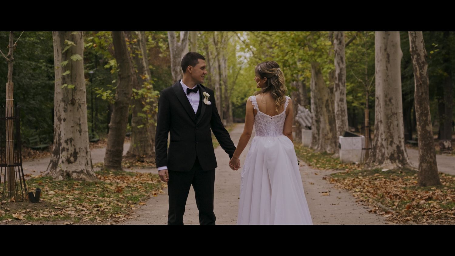A romantic wedding portrait of a couple walking through a tree-lined path in formal attire.