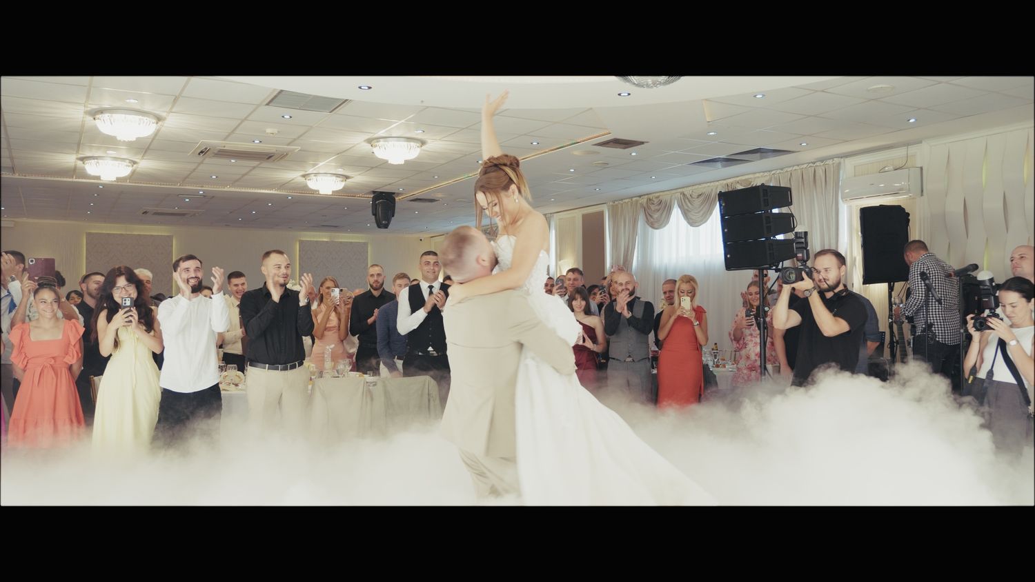 Couple performs their first dance in a grand ballroom filled with wedding guests and atmospheric fog.