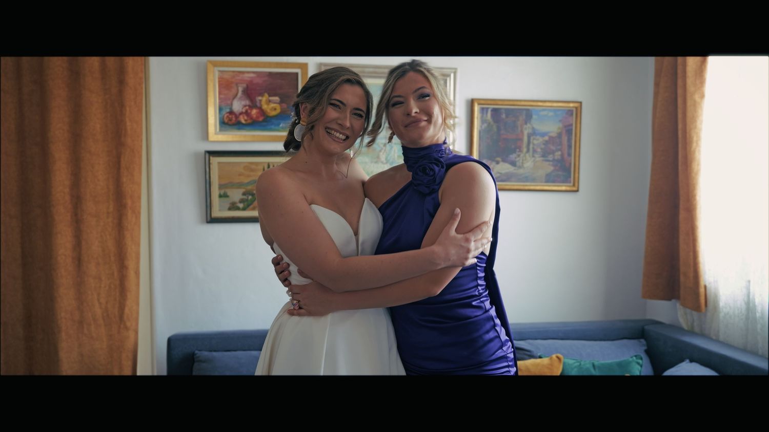 Wedding party members embrace in a sunlit room with artwork on the walls.