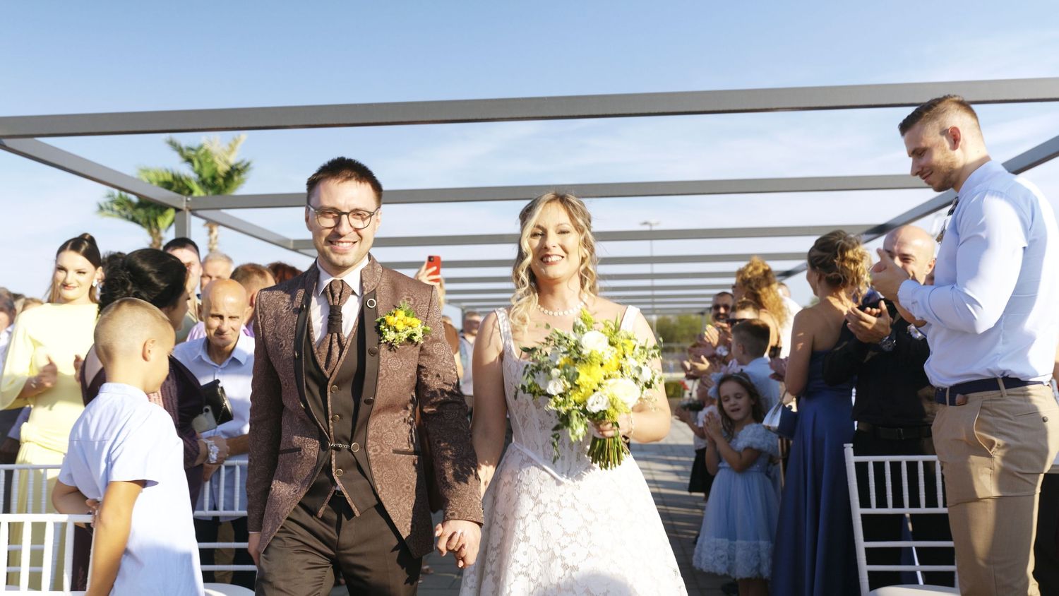 Wedding ceremony under white pergola with guests seated in outdoor venue.