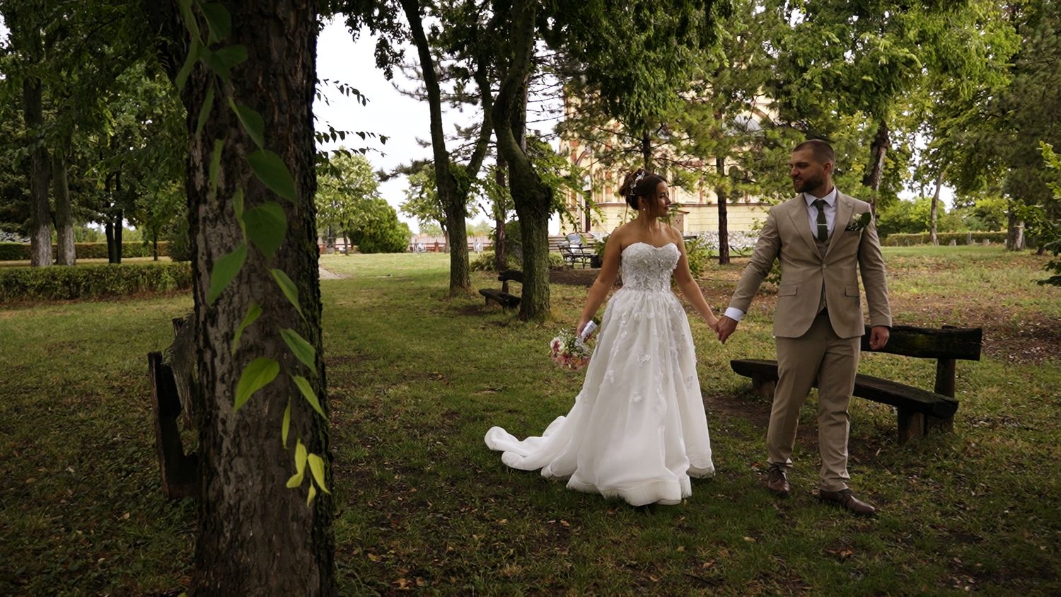 Wedding couple strolling through park in elegant attire on sunny day.