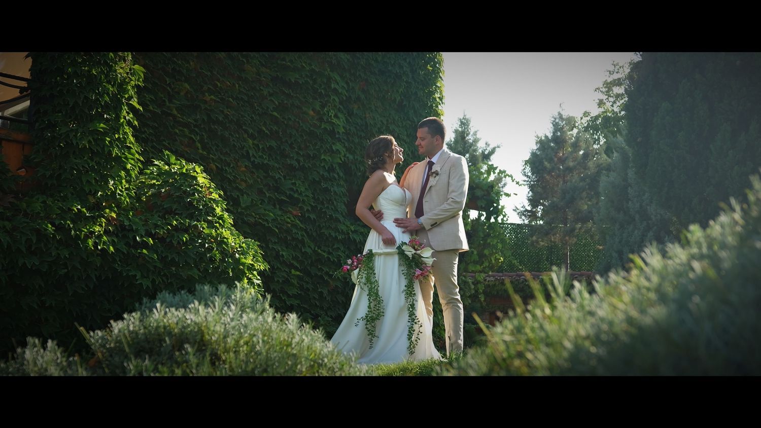 A couple stands together in a garden setting with lush greenery and sunlight.